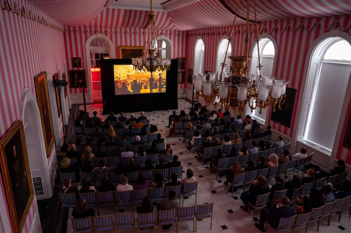 A large crowd of people sit in the Tent Room at Rideau Hall and watch a movie on a large screen