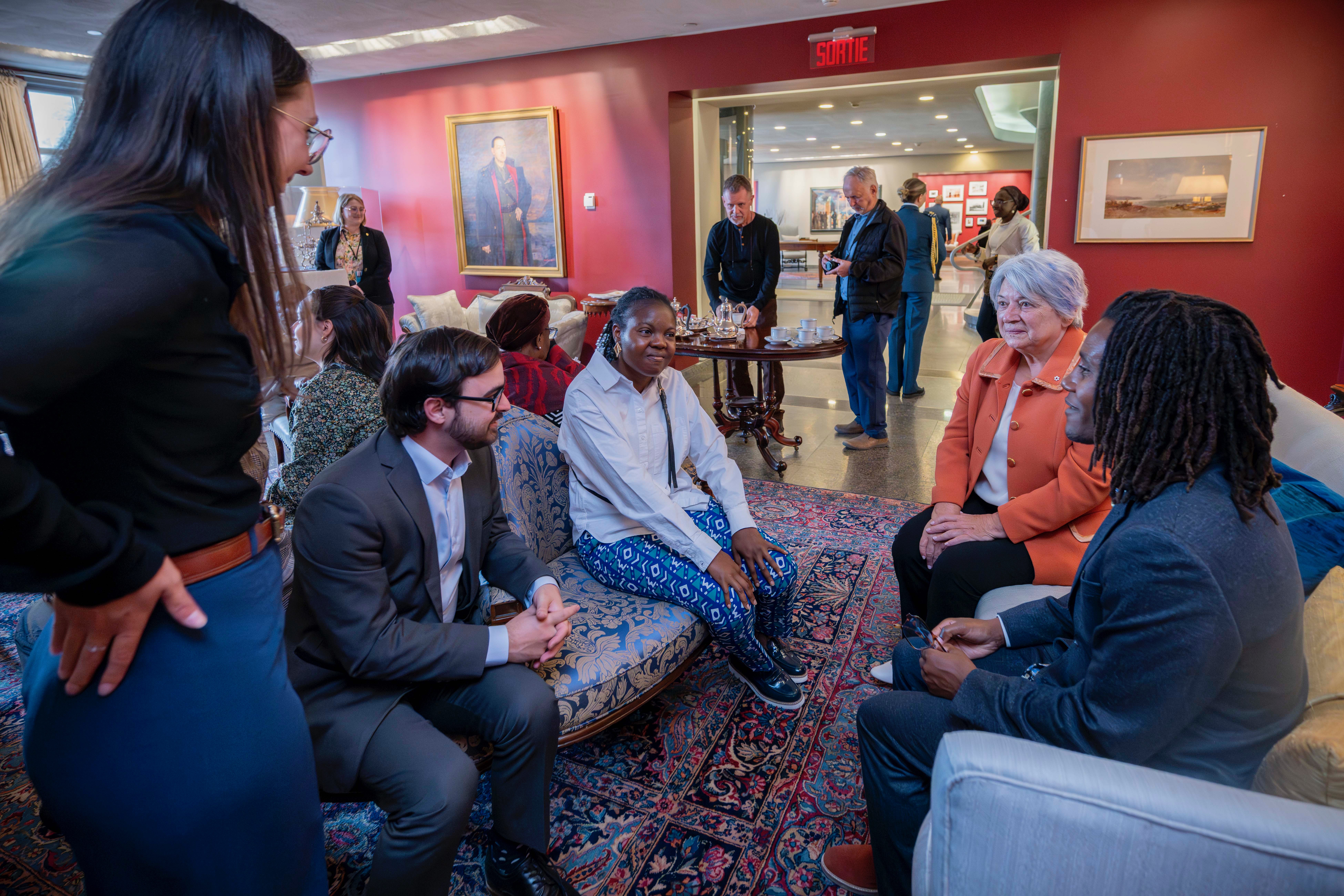 A group of people gathered in a warmly lit room, seated on patterned sofas. Governor General Mary Simon is engaged in conversation. A few people stand in the background by a table with tea and coffee.