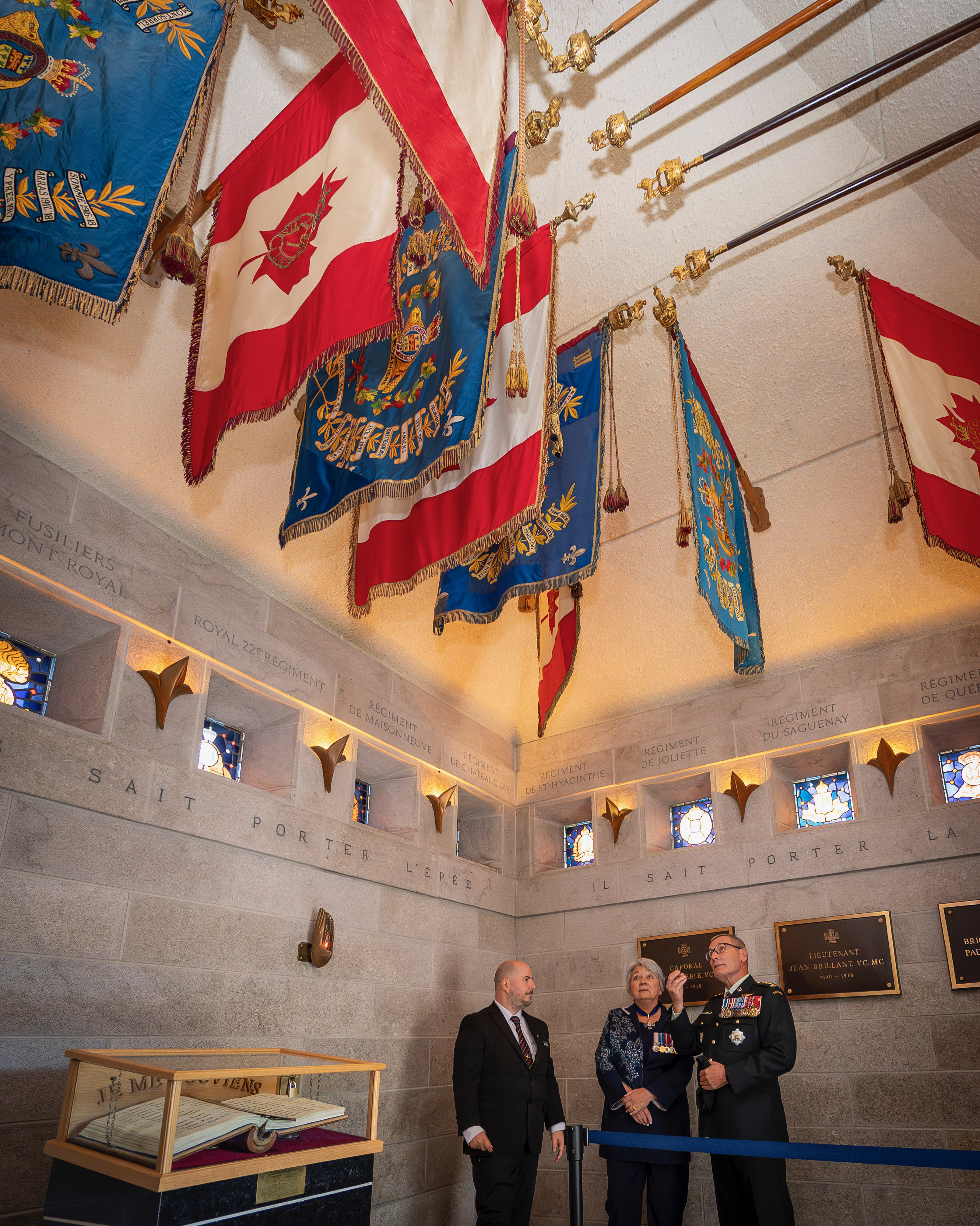 Governor General Mary Simon standing with two people beneath colorful, ornate flags hanging from the ceiling in a solemn room. Plaques adorn the walls.