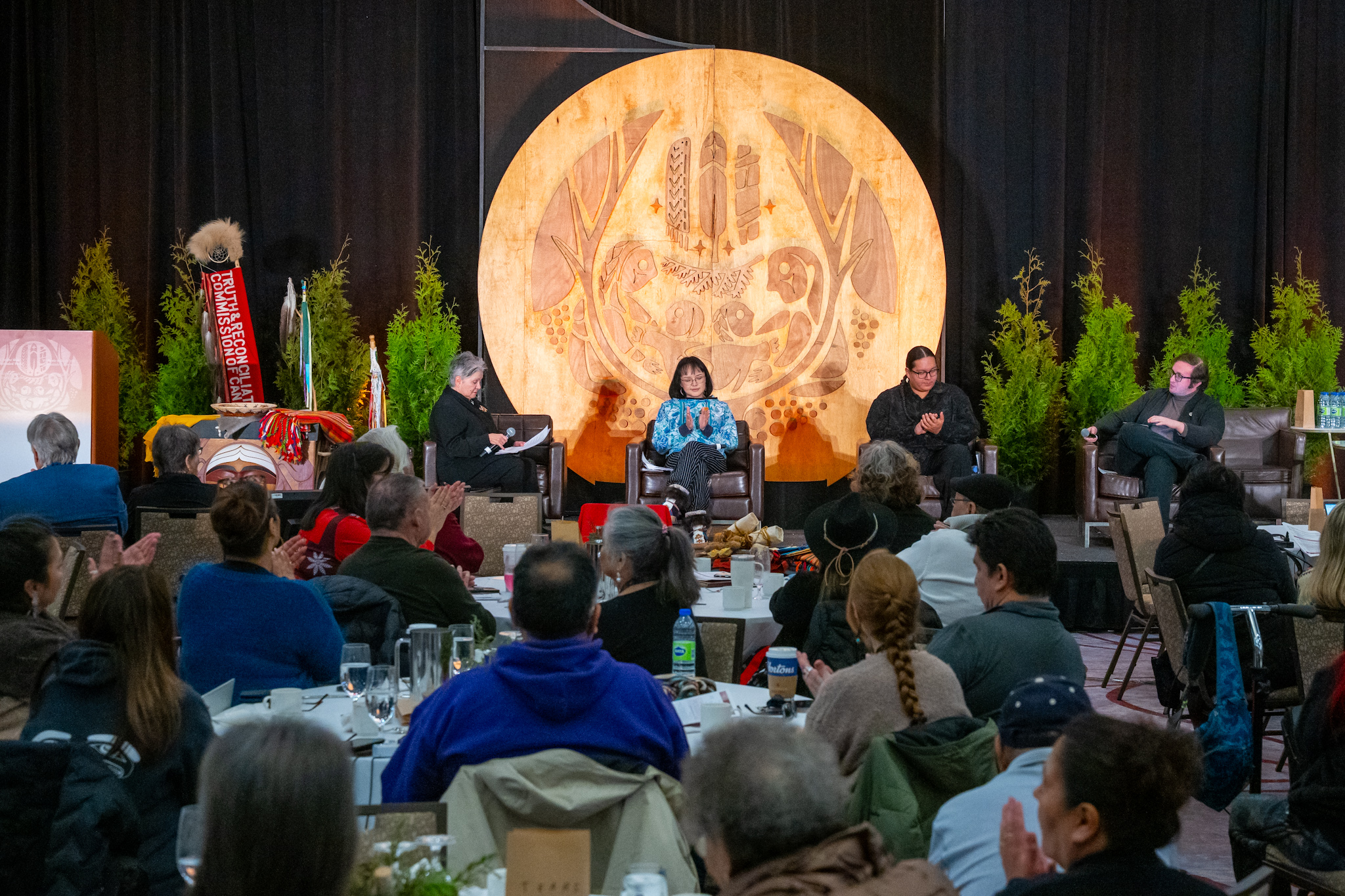 A panel of people sitting on a stage. An audience is seated at round tables, clapping.