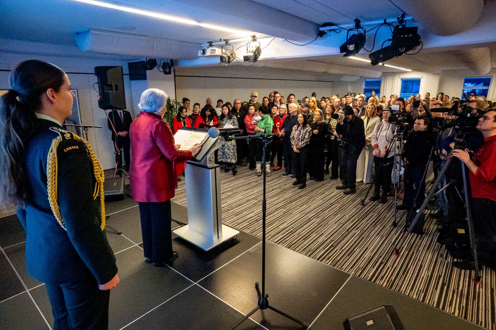 Governor General Mary Simon speaking at a podium to a group of people who are standing. Photo is taken from behind the Governor General. 