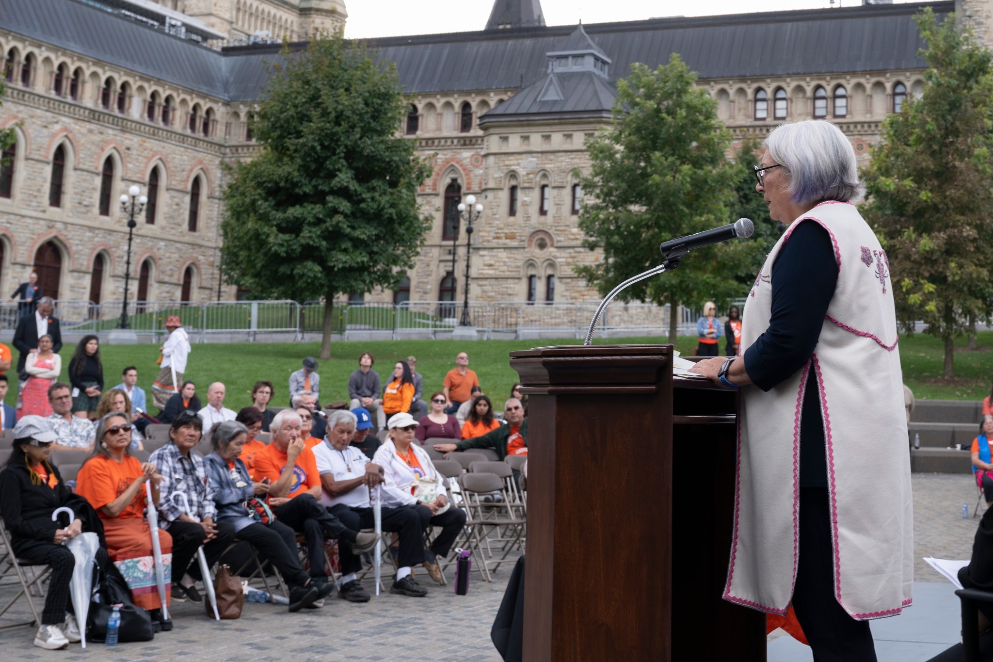 Governor General Mary Simon is standing at a podium. People are seated in front of her. Many people are wearing orange shirts. There is a large stone building in a background.