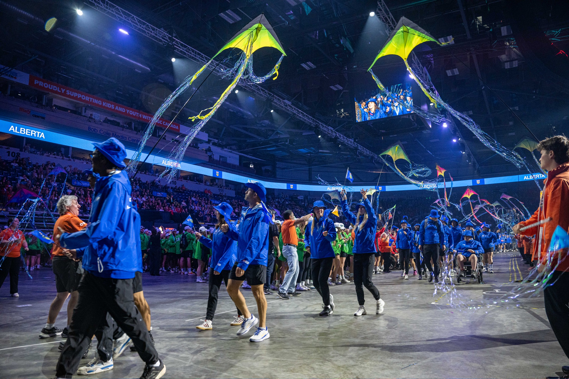 Athletes walking in a large stadium. Large kites fly above. 