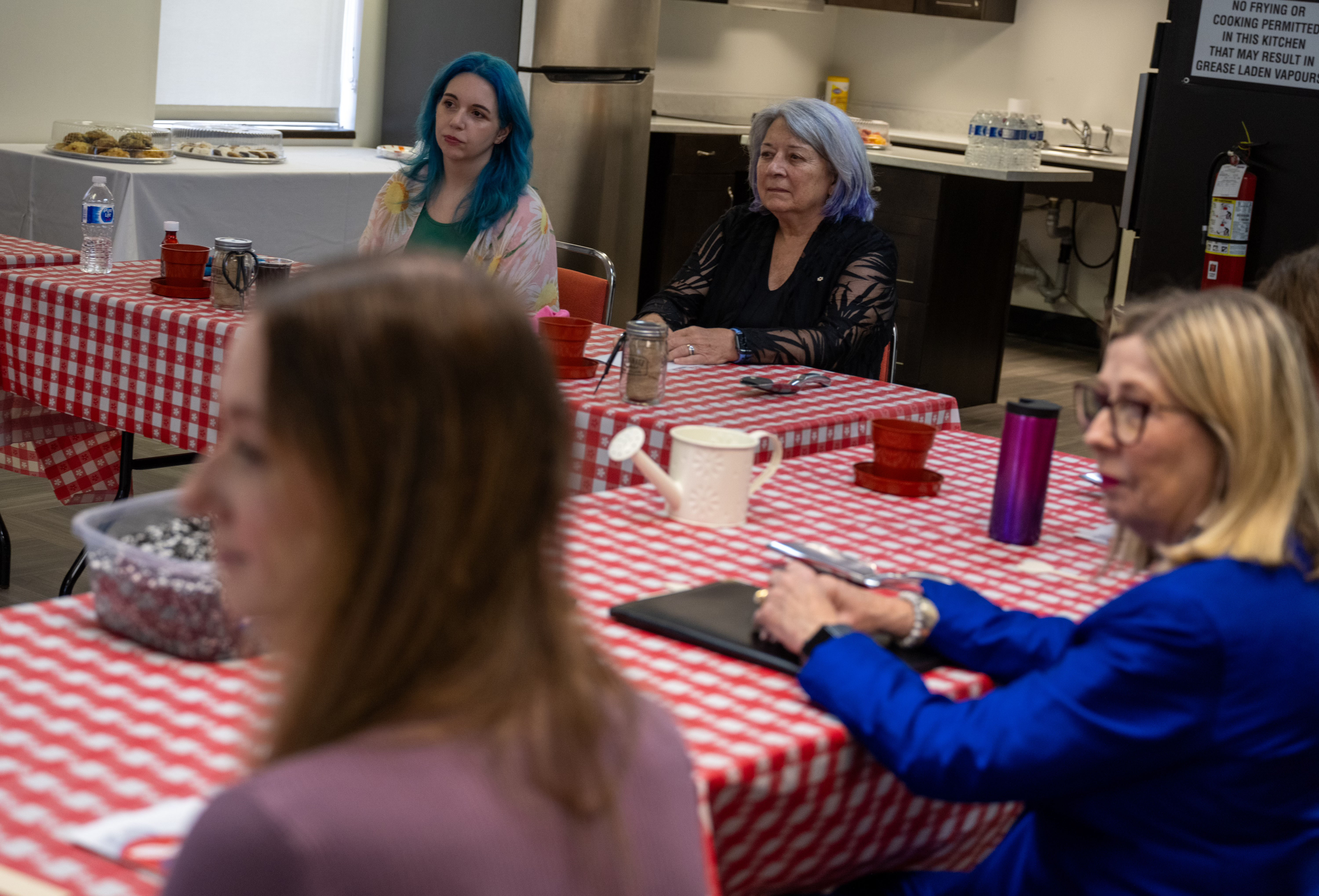 A group of people sit attentively around a table with a red-checkered tablecloth in a kitchen setting.