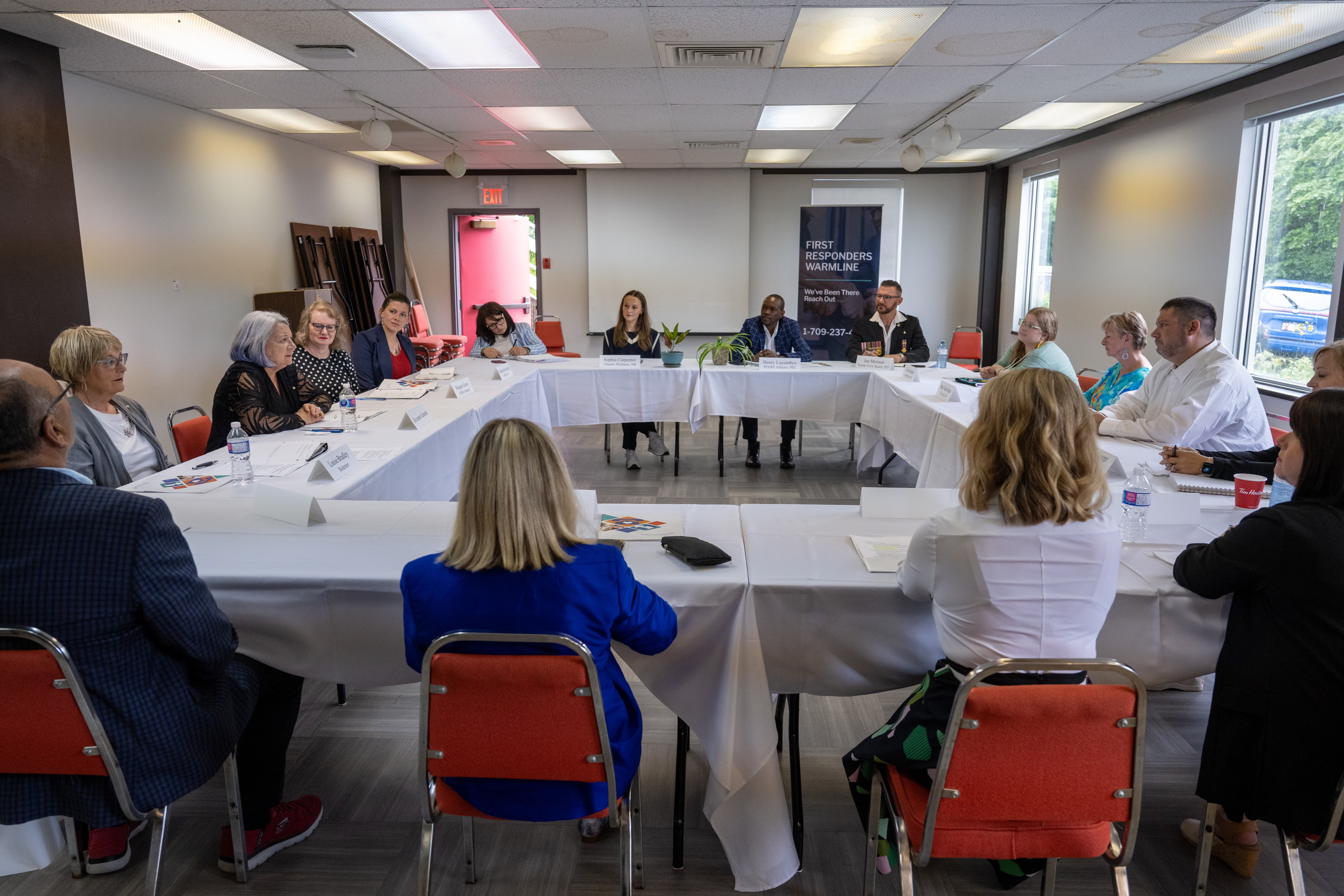 A diverse group of people sit around a U-shaped table in a meeting room. The atmosphere is professional and collaborative, with papers and nameplates on the table.