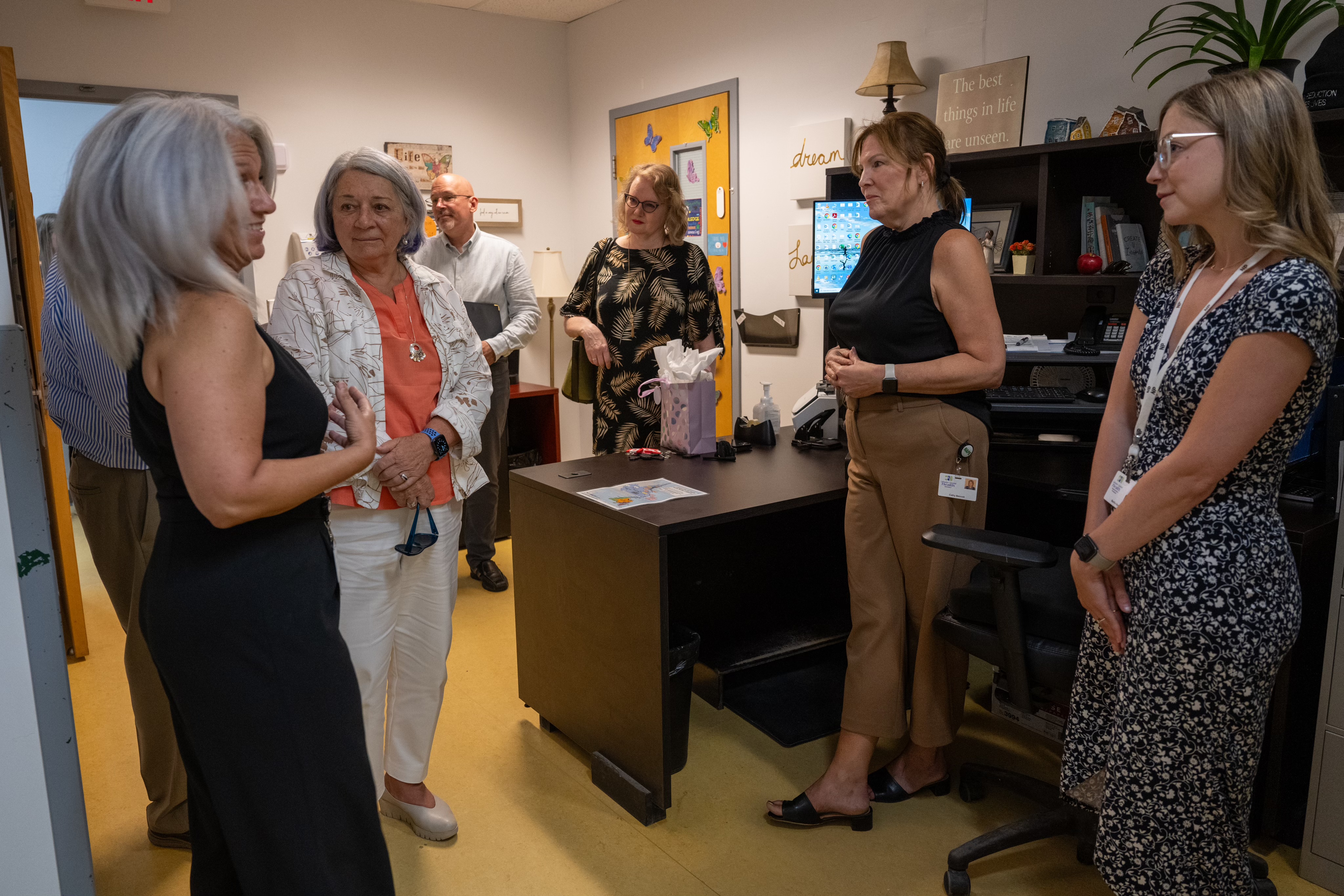 A group of five people converse in an office, surrounded by shelves and a desk.