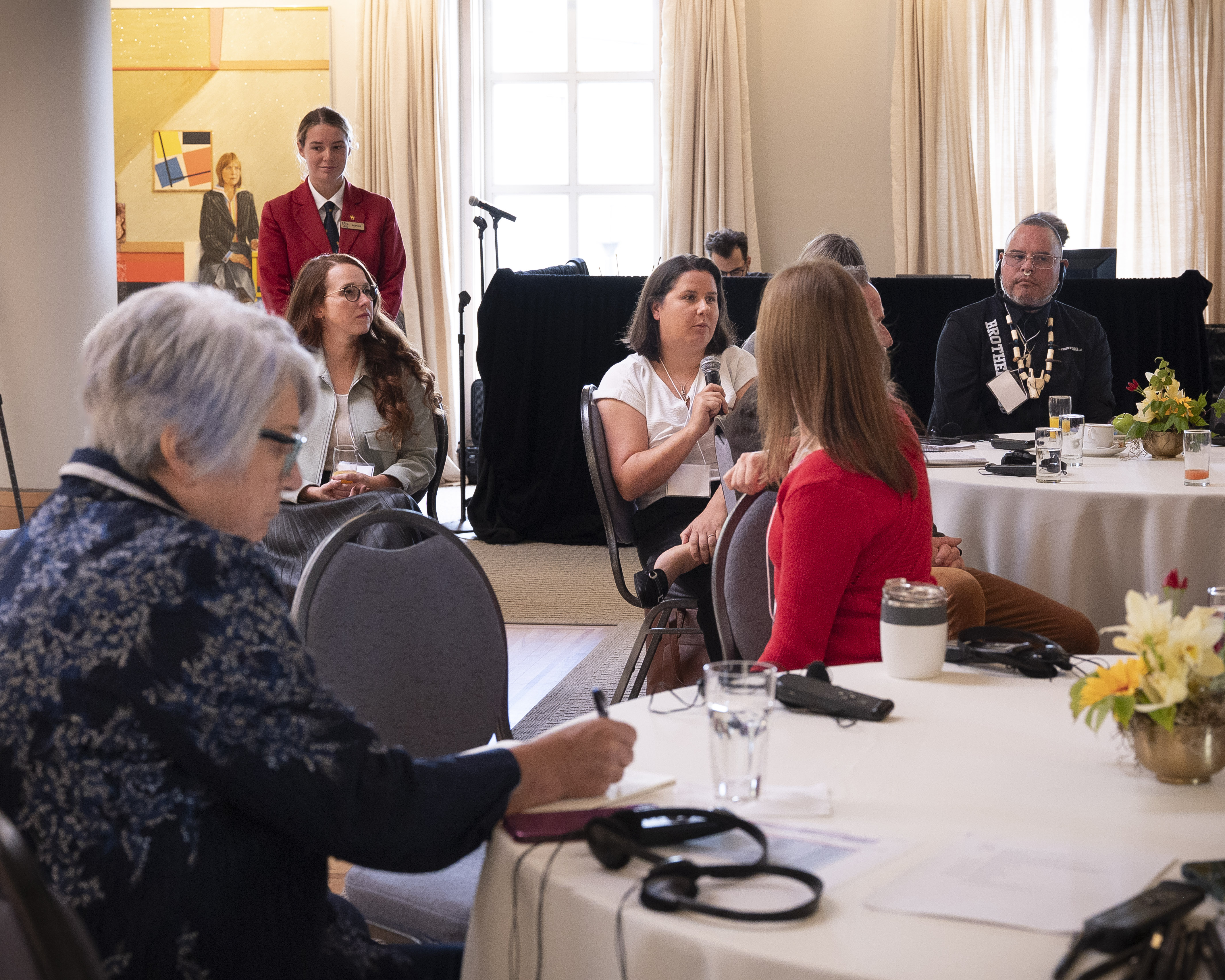A group of people sitting at round tables. One person is holding a microphone.