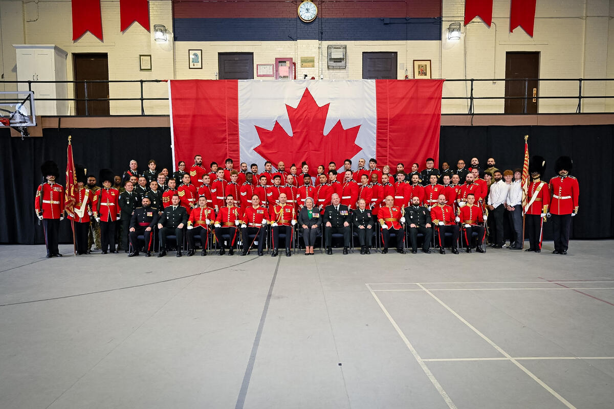 Governor General Mary Simon and a large group of military personnel and civilians posing for a group photo in front of a Canadian flag. The group includes soldiers seated and standing in rows in a large gymnasium.