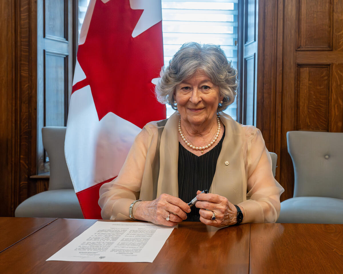 Governor General Arbour holding a pen. A piece of paper is in front of her on a large wooden table. There is a large Canada flag behind her.