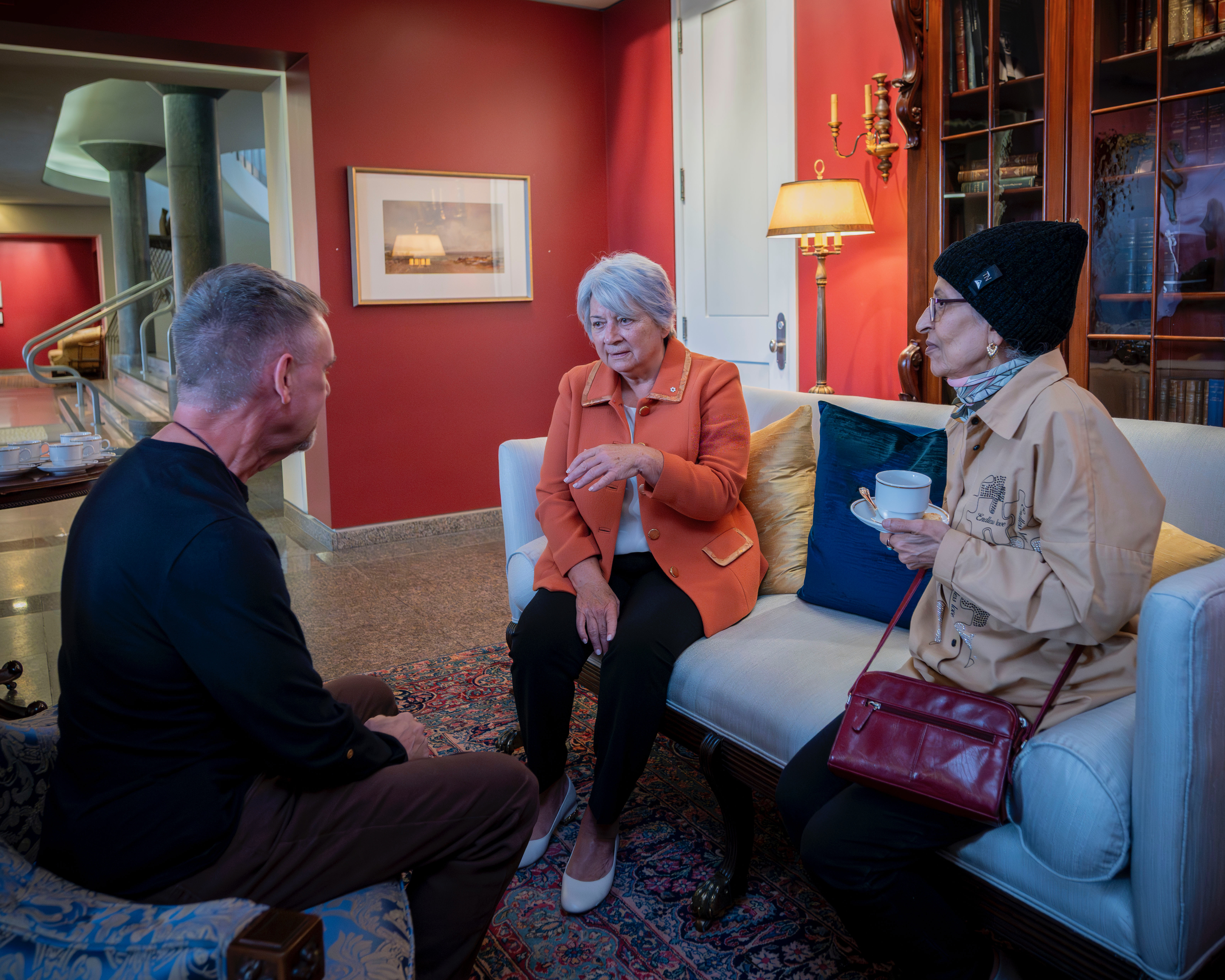 A group of people gathered in a warmly lit room, seated on patterned sofas. Governor General Mary Simon is engaged in conversation. A few people stand in the background by a table with tea and coffee.