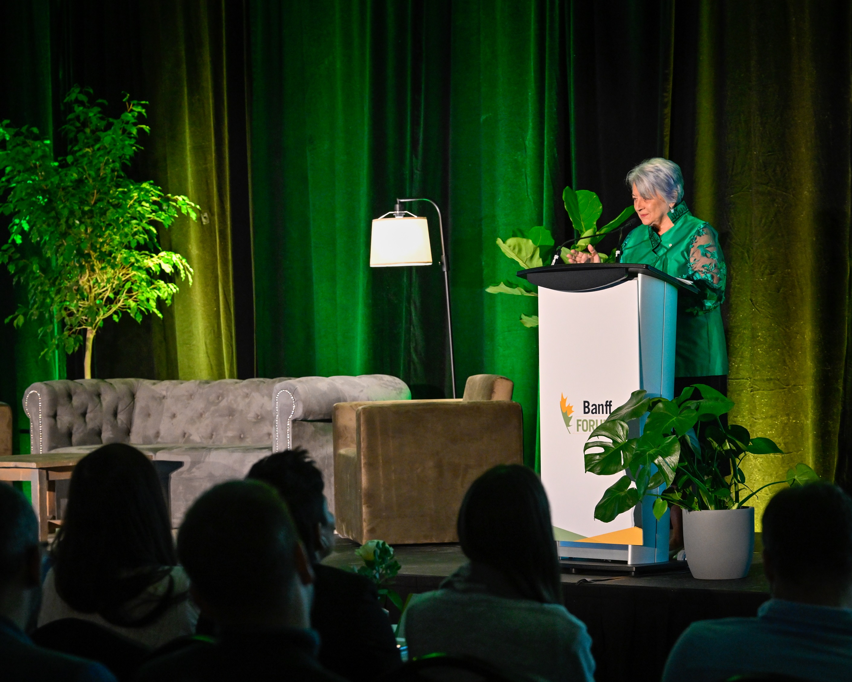 Governor General Mary Simon speaking at a podium with "Banff Forum" branding. She's on a stage with green curtains, a sofa, plants, and a lamp.