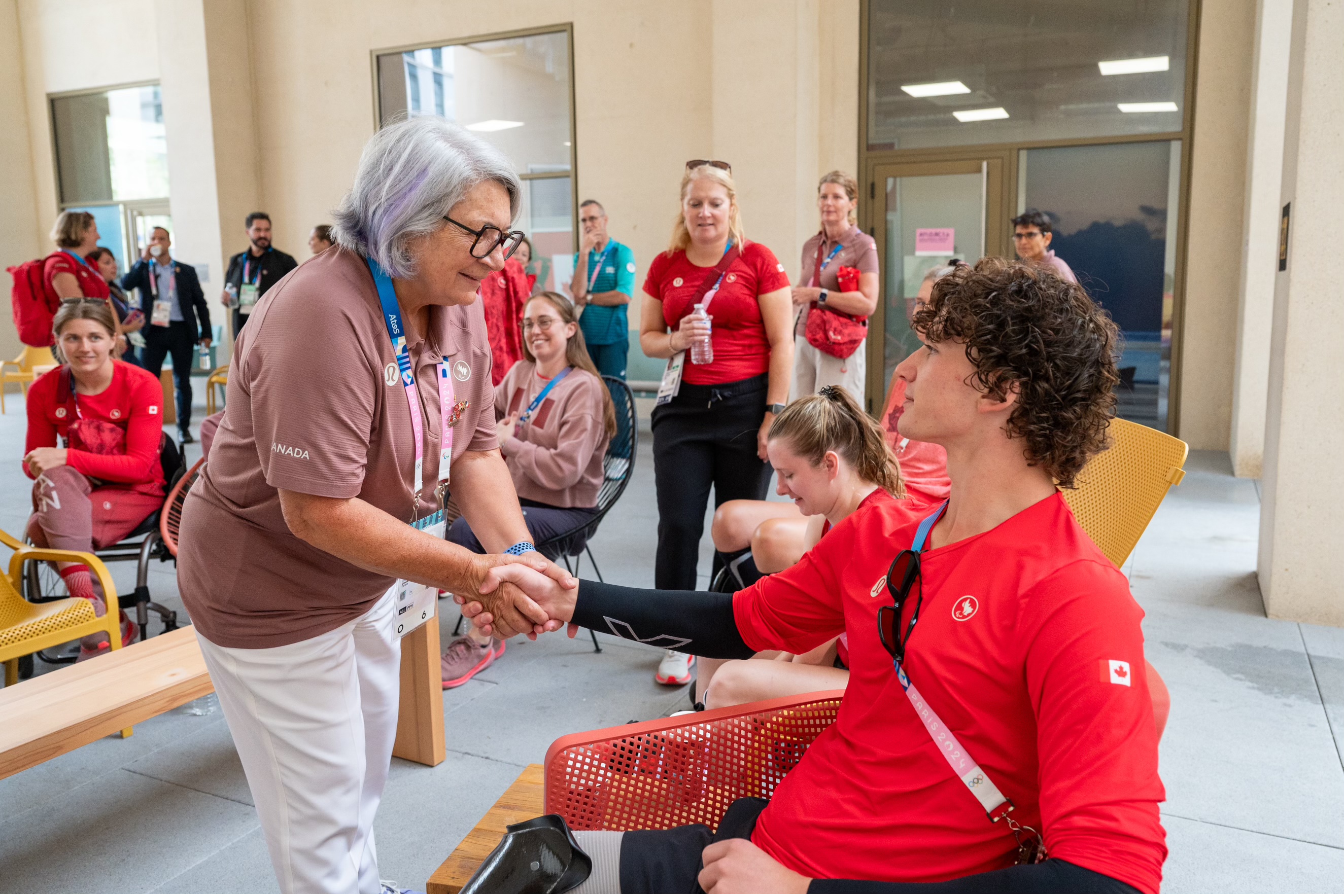 La gouverneure générale Mary Simon se penche en avant pour serrer la main d'un athlète d'Équipe Canada assis. Plusieurs personnes, dont d'autres athlètes et officiels, sourient et observent l'interaction.