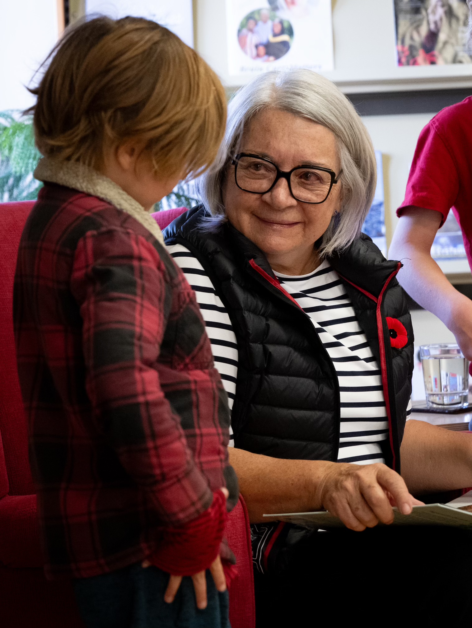 Governor General Mary Simon sits in a red chair, while holding a book and smiling at child who is standing beside her.