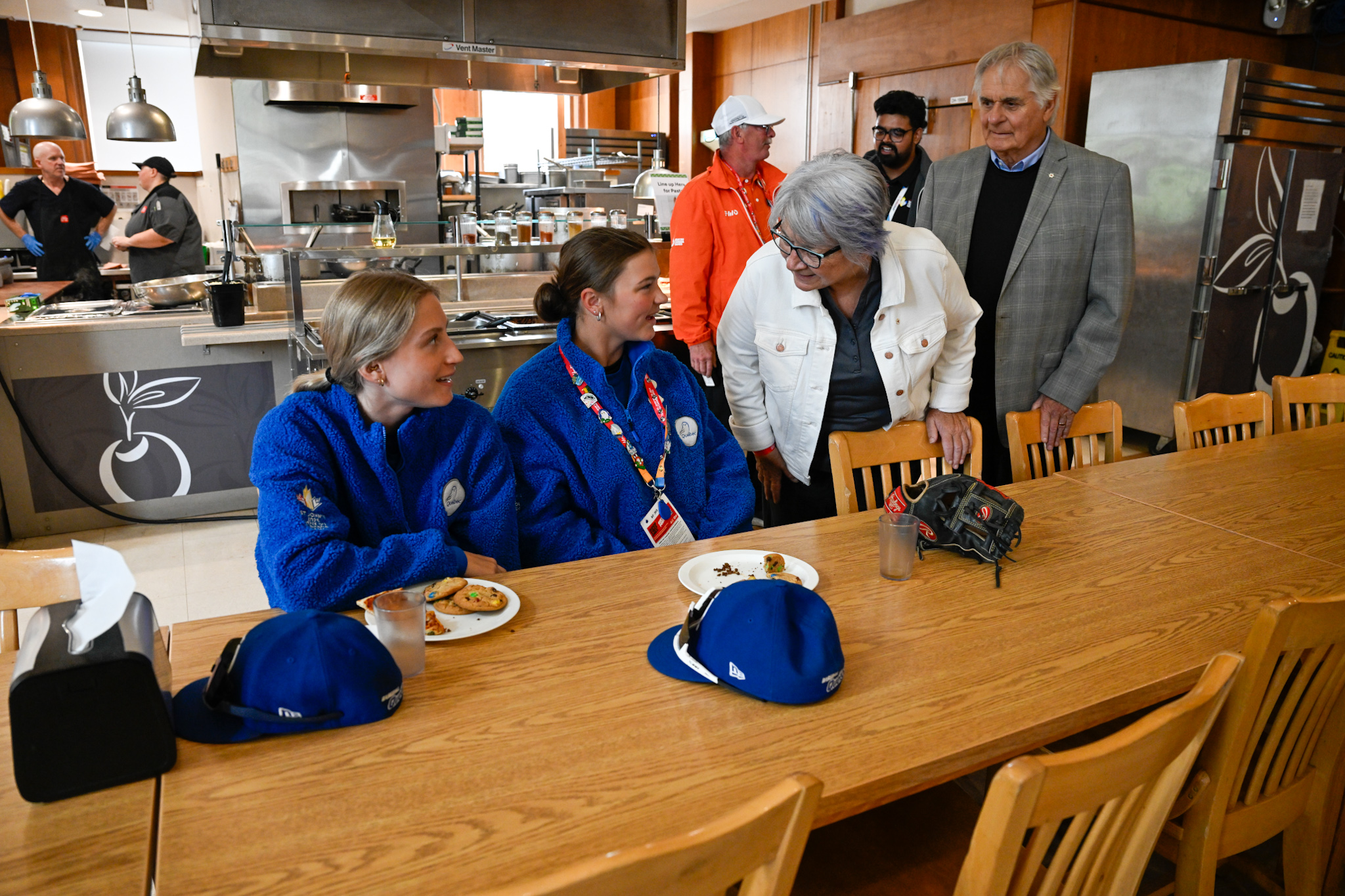 Governor General Mary Simon and Mr. Whit Fraser speaking to two young people dressed in blue fleece zip-ups who are sitting at a long table with plates in front of them.
