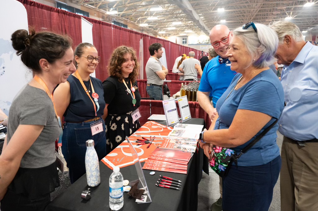 La gouverneure générale Mary Simon s'entretient avec des vendeuses qui se tiennent à un stand. Des stylos et des brochures sont posés sur la table.
