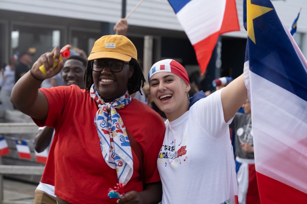 Deux personnes sourient pour une photo. Elles participent aux festivités de la Fête nationale de l'Acadie. L'une d'elles tient un drapeau acadien. L'autre tient un mini klaxon.