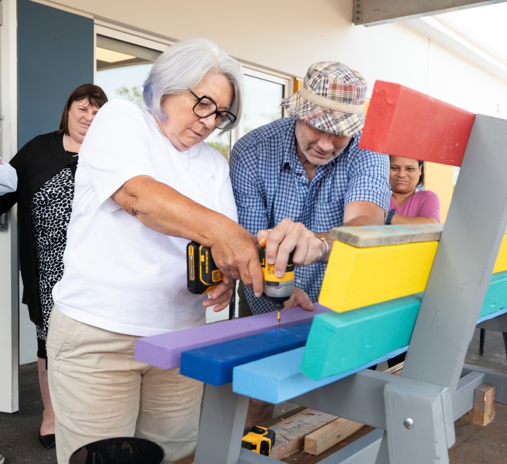 La gouverneure générale Mary Simon utilise une perceuse sur un banc aux couleurs de l'arc-en-ciel pendant qu'une personne l'aide à tenir la perceuse.