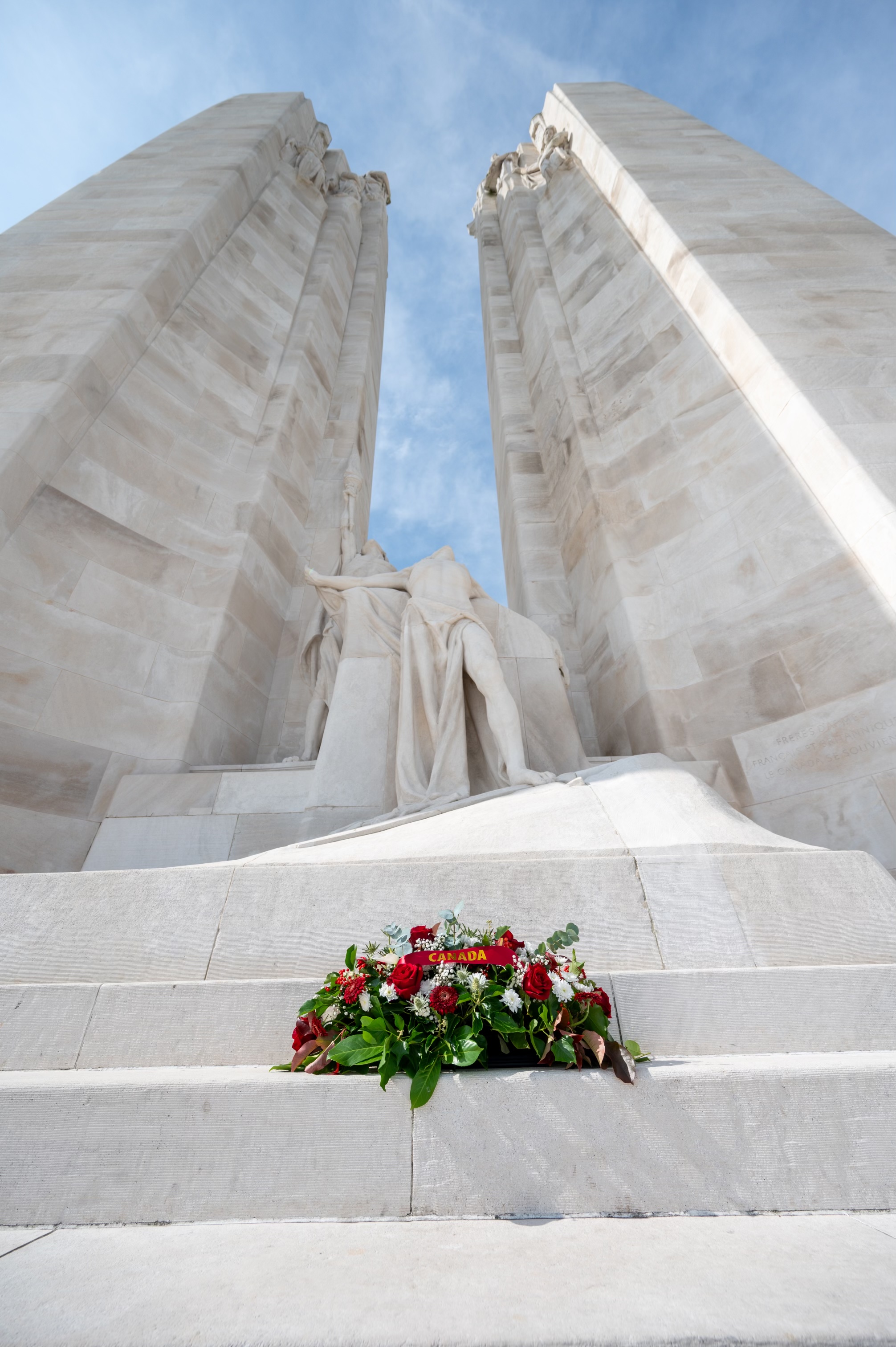 Une couronne de fleurs rouges et blanches avec des feuilles vertes repose sur les marches devant un grand monument en pierre blanche avec deux hautes colonnnes et une figure sculptée.