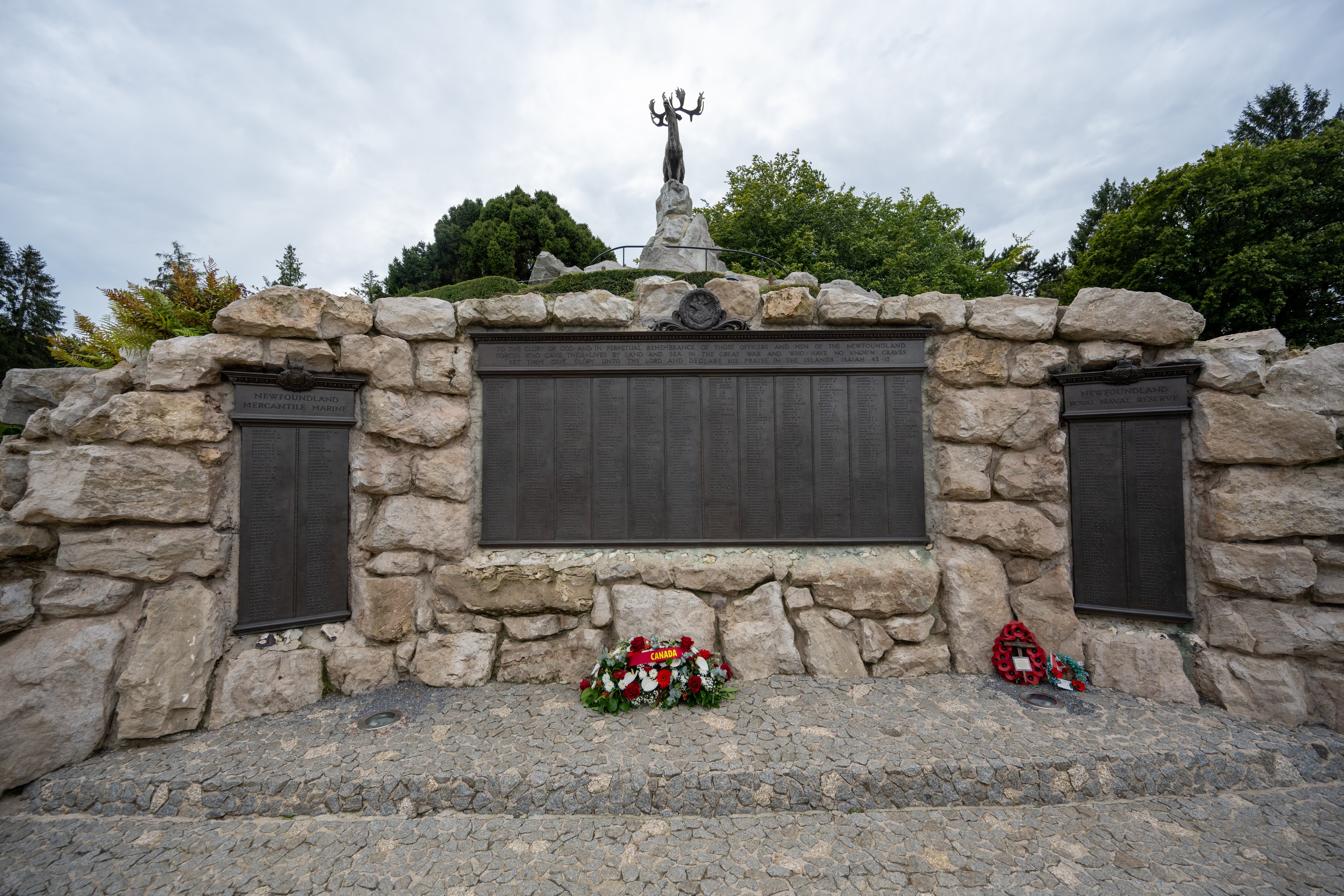Un mémorial composé de grands blocs de pierre, avec des plaques portant des noms et ornées de couronnes et de hommages floraux à sa base. Au sommet, une statue représentant une figure.