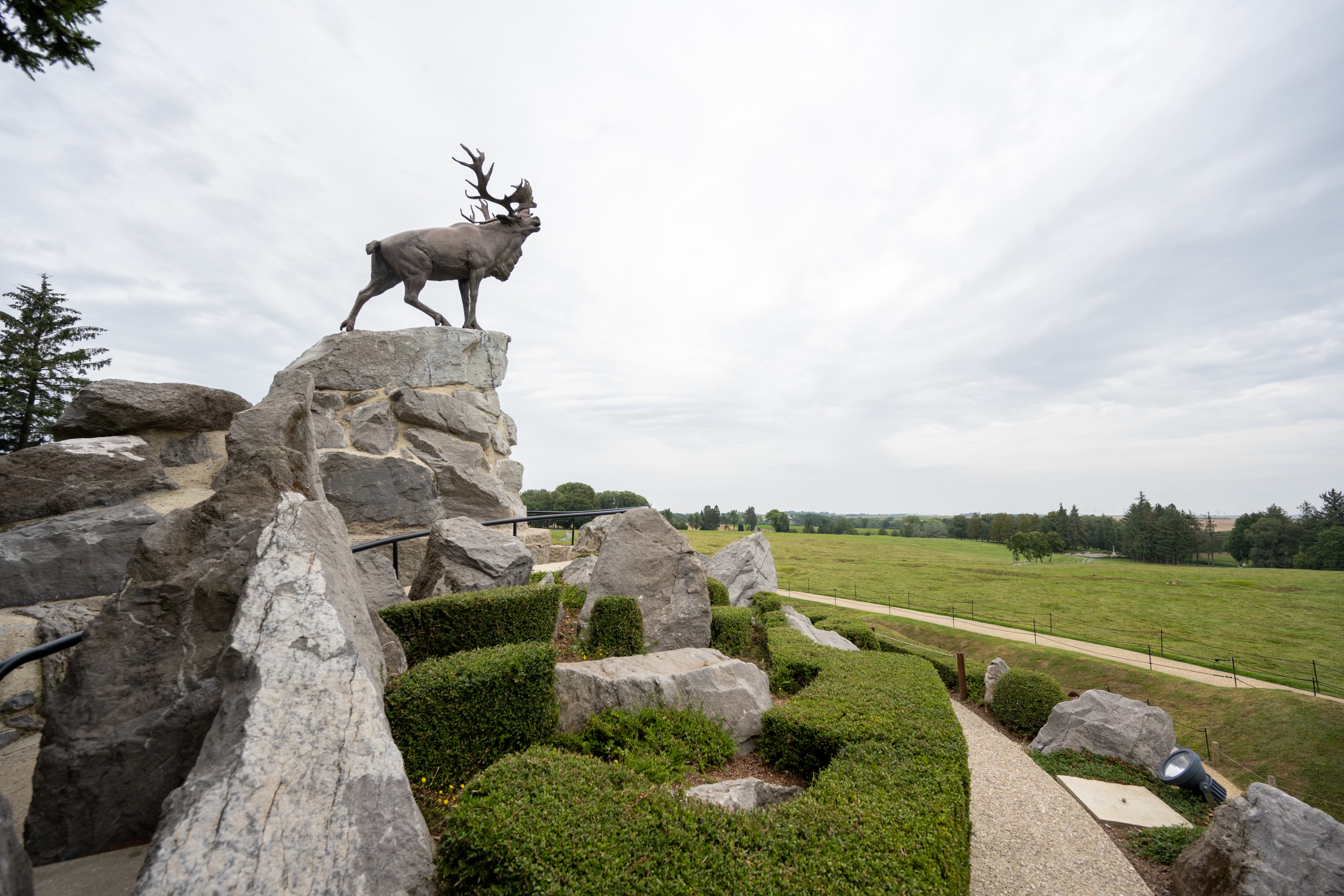 Une statue en bronze de caribou se dresse au sommet d'une formation rocheuse, entourée de buis taillés.