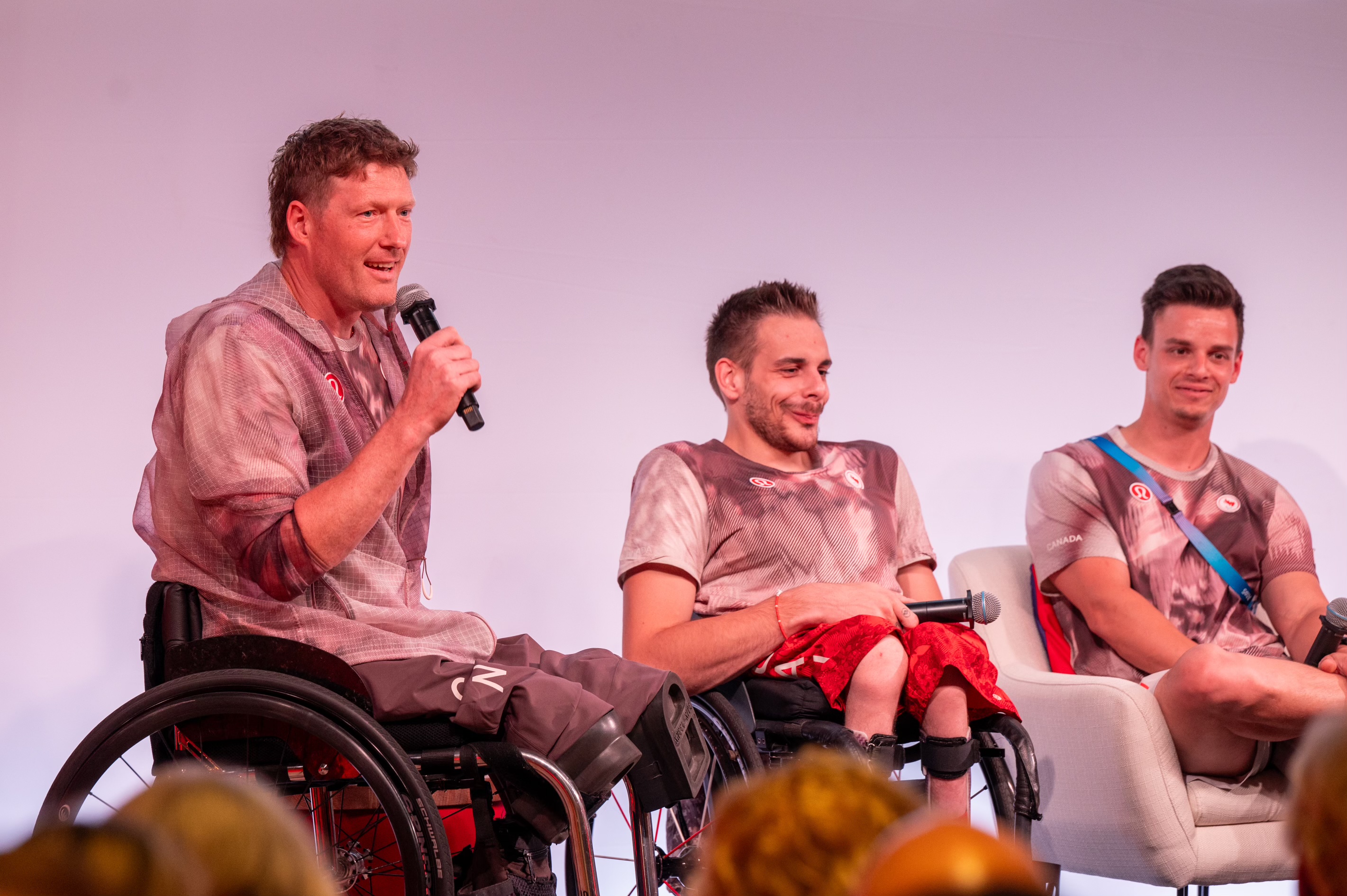 Trois personnes, dont deux en fauteuils roulants, participent à une discussion. La personne à gauche parle dans un microphone. Un public est visible au premier plan.