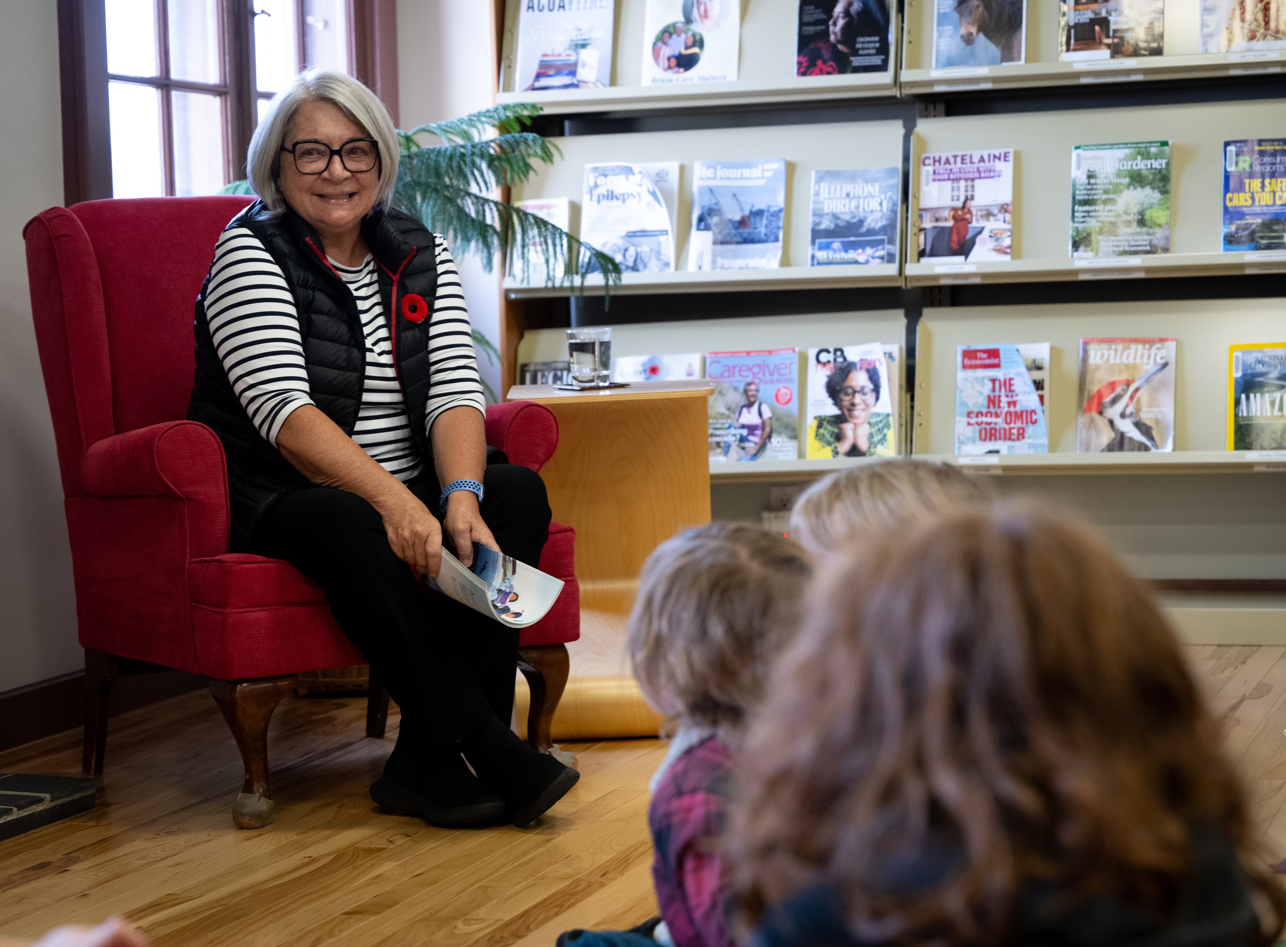Governor General Mary Simon sits in a red chair, smiling, and holding a book while talking to a small group of children.