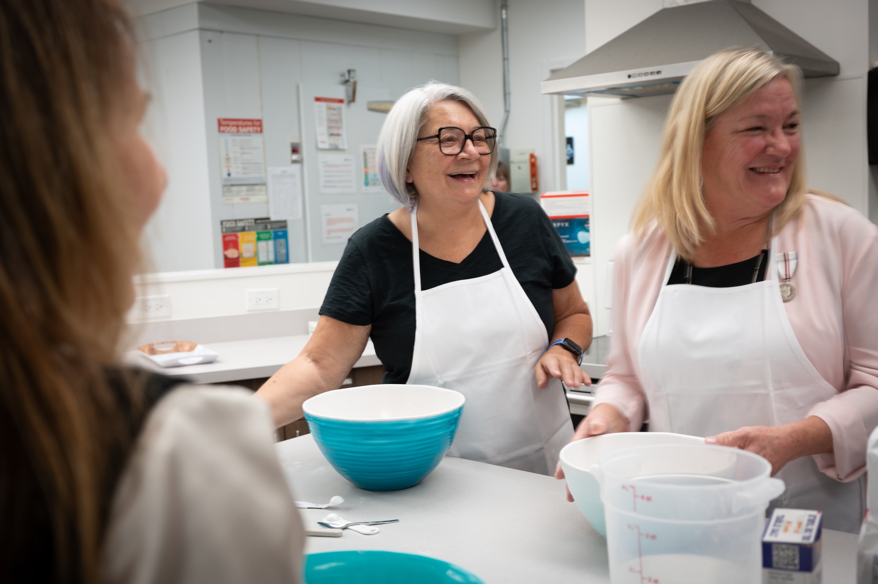 Governor General Mary Simon and another person, wearing white aprons, smile and chat in the kitchen as they prepare ingredients in large mixing bowls.