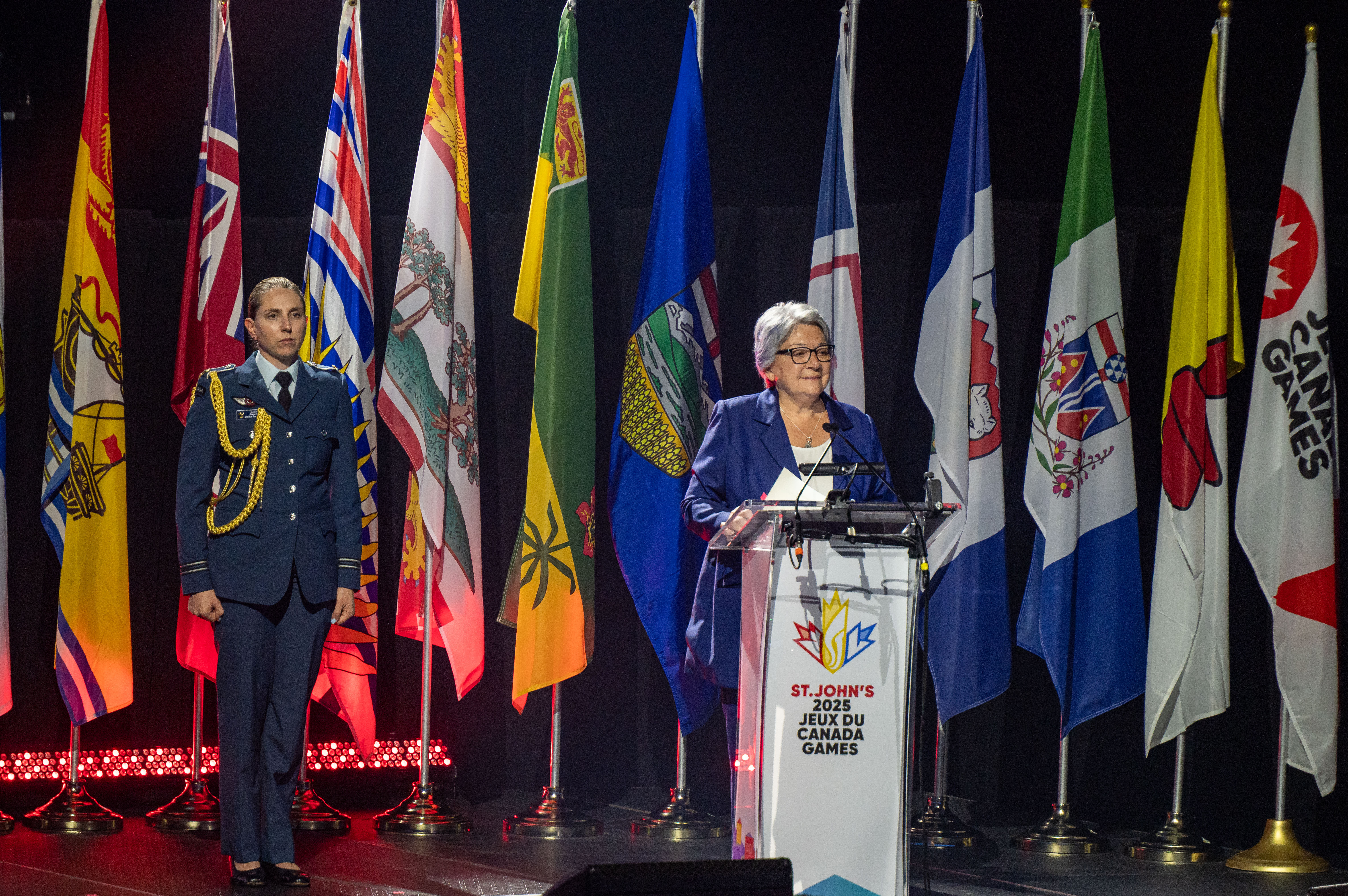 Governor General Mary Simon standing at a podium. Provincial flags are behind her. A woman in military uniform is standing to her right.
