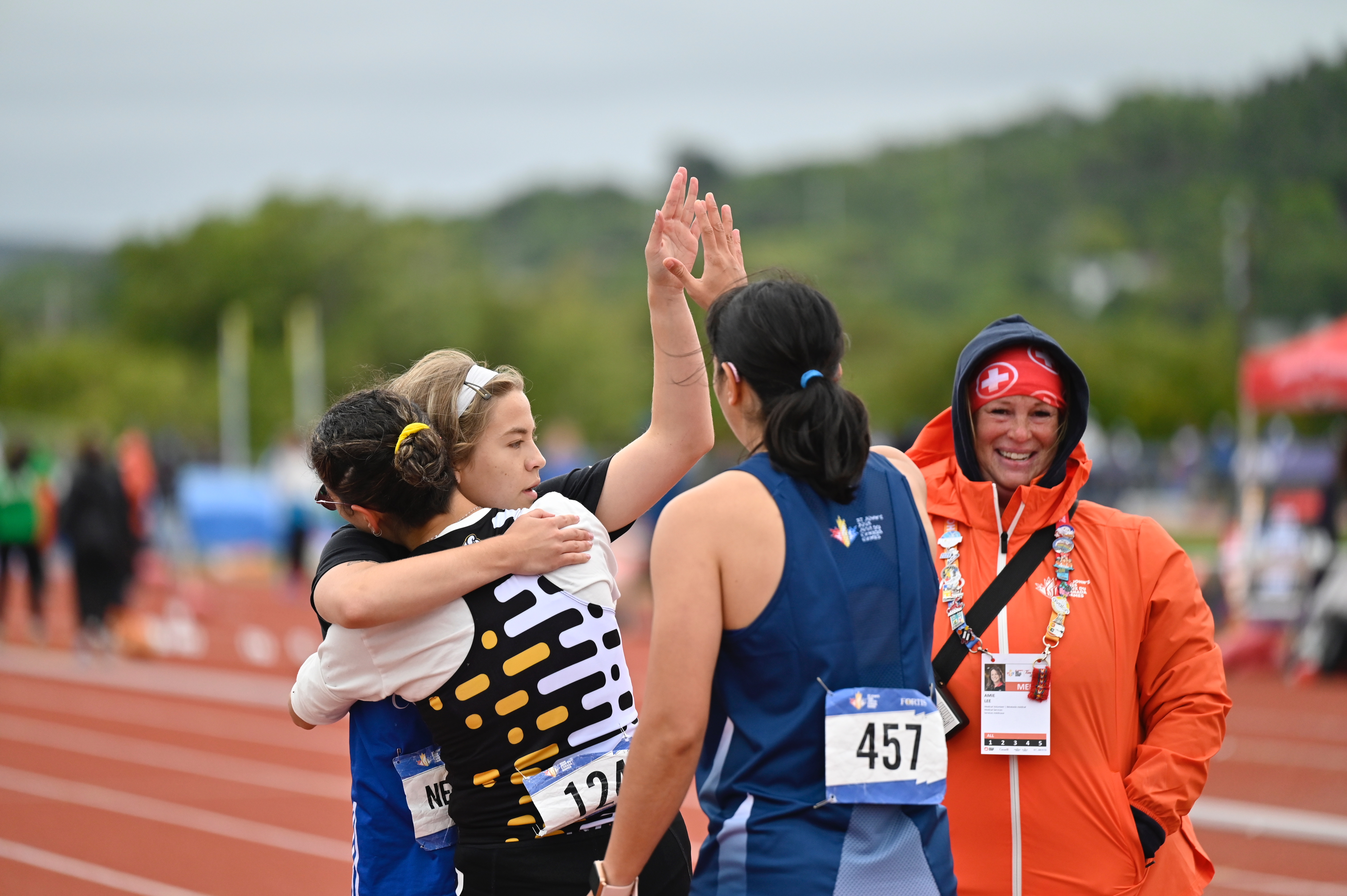 Athletes hugging and high-fiving on the running track. A First Aid responder in an orange coat is standing near them.