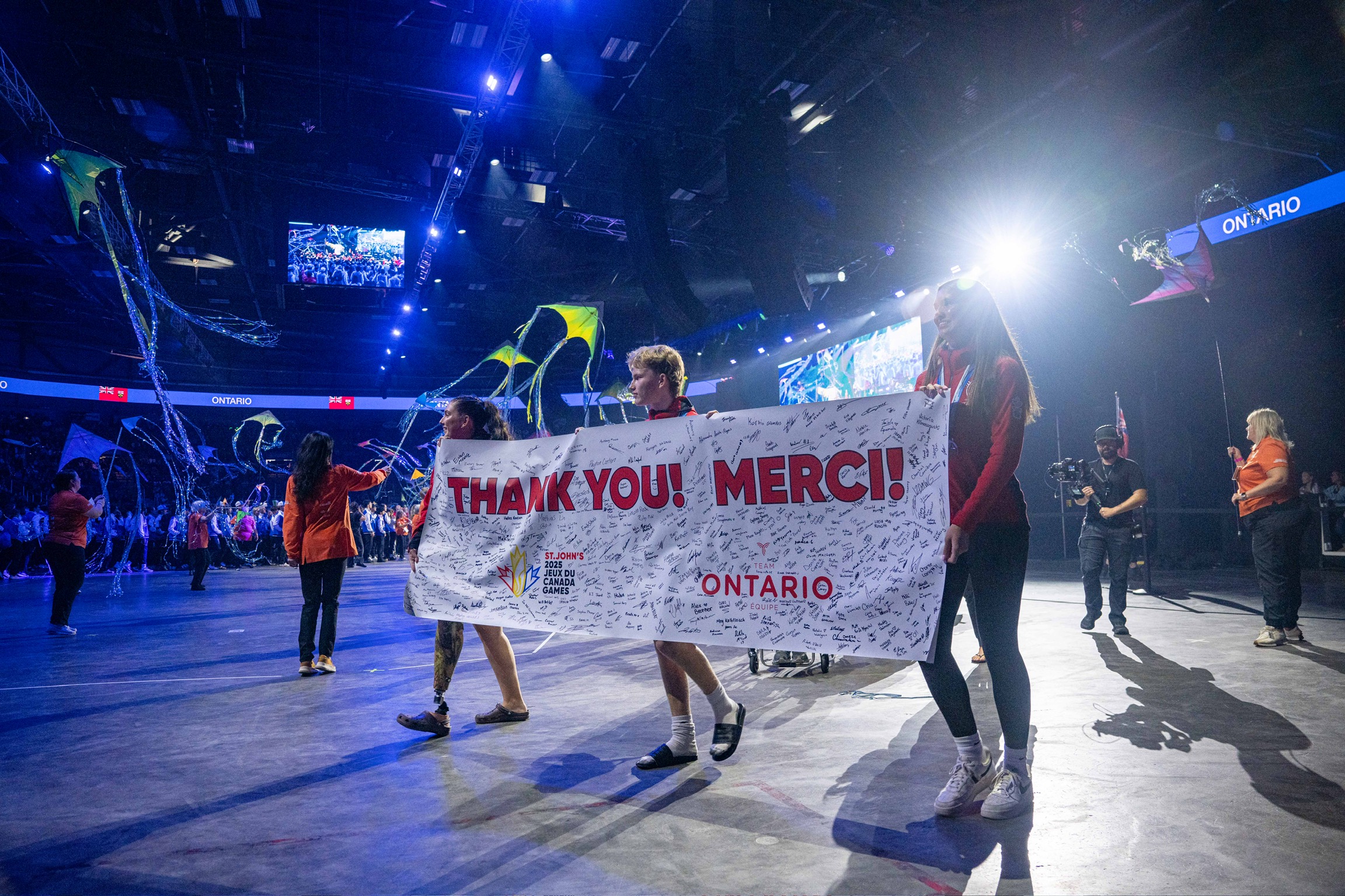 Athletes walking in a parade in a large stadium holding a banner that has "Thank you! Merci!" written on it, filled with signatures.