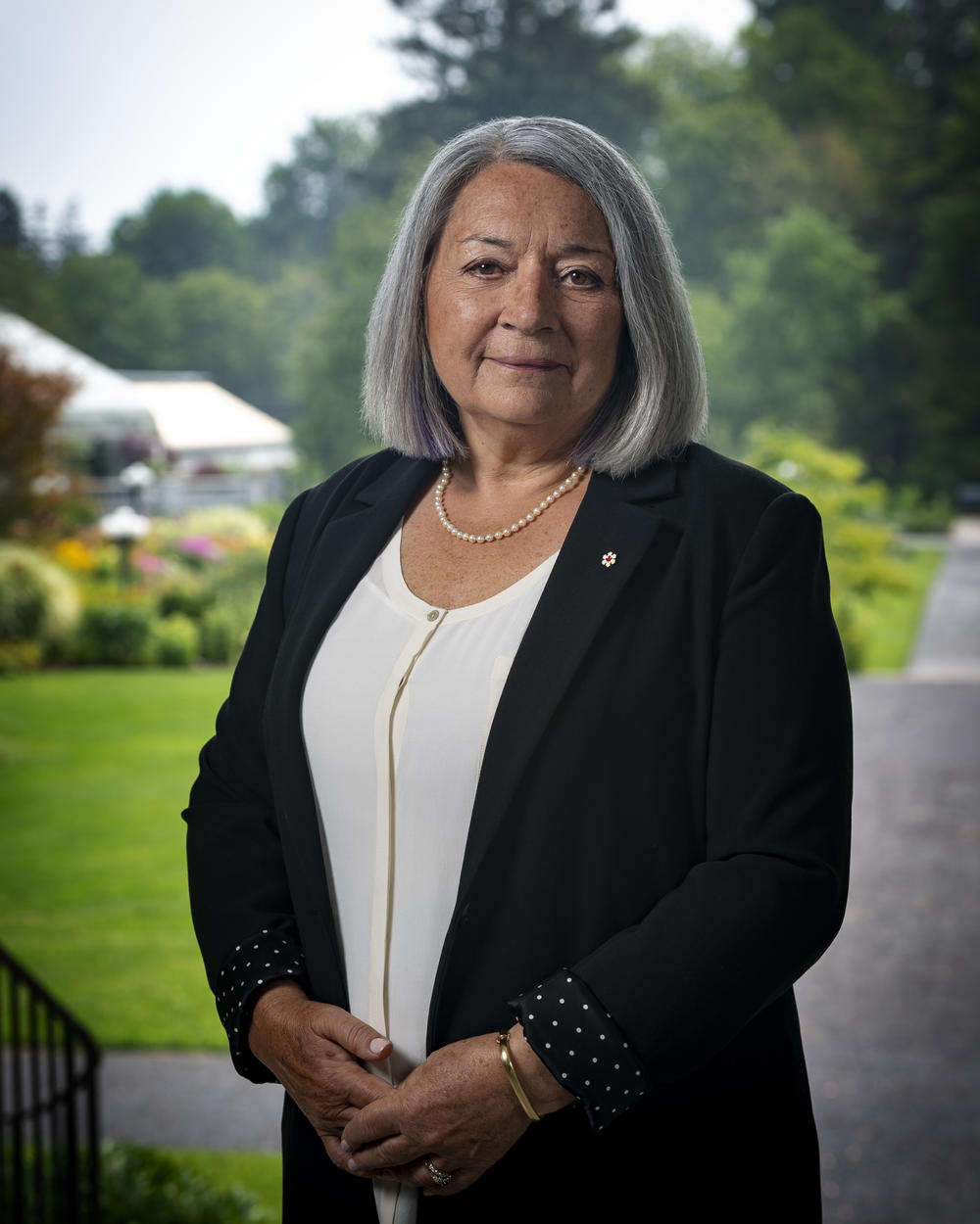 Photo officielle de la gouverneure générale Mary Simon. Elle est à l'extérieur. Les jardins de Rideau Hall sont visibles à l'arrière-plan.