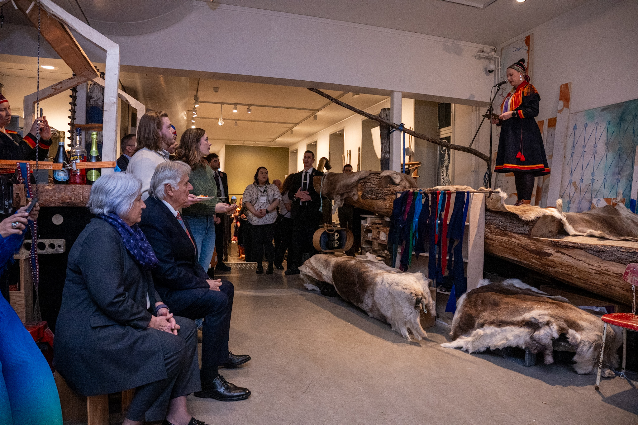 A woman in traditional Sámi attire speaks to a seated audience in a room decorated with animal pelts, textiles, and wooden beams. Governor General Mary Simon and Mr. Whit Fraser are seated, listening.