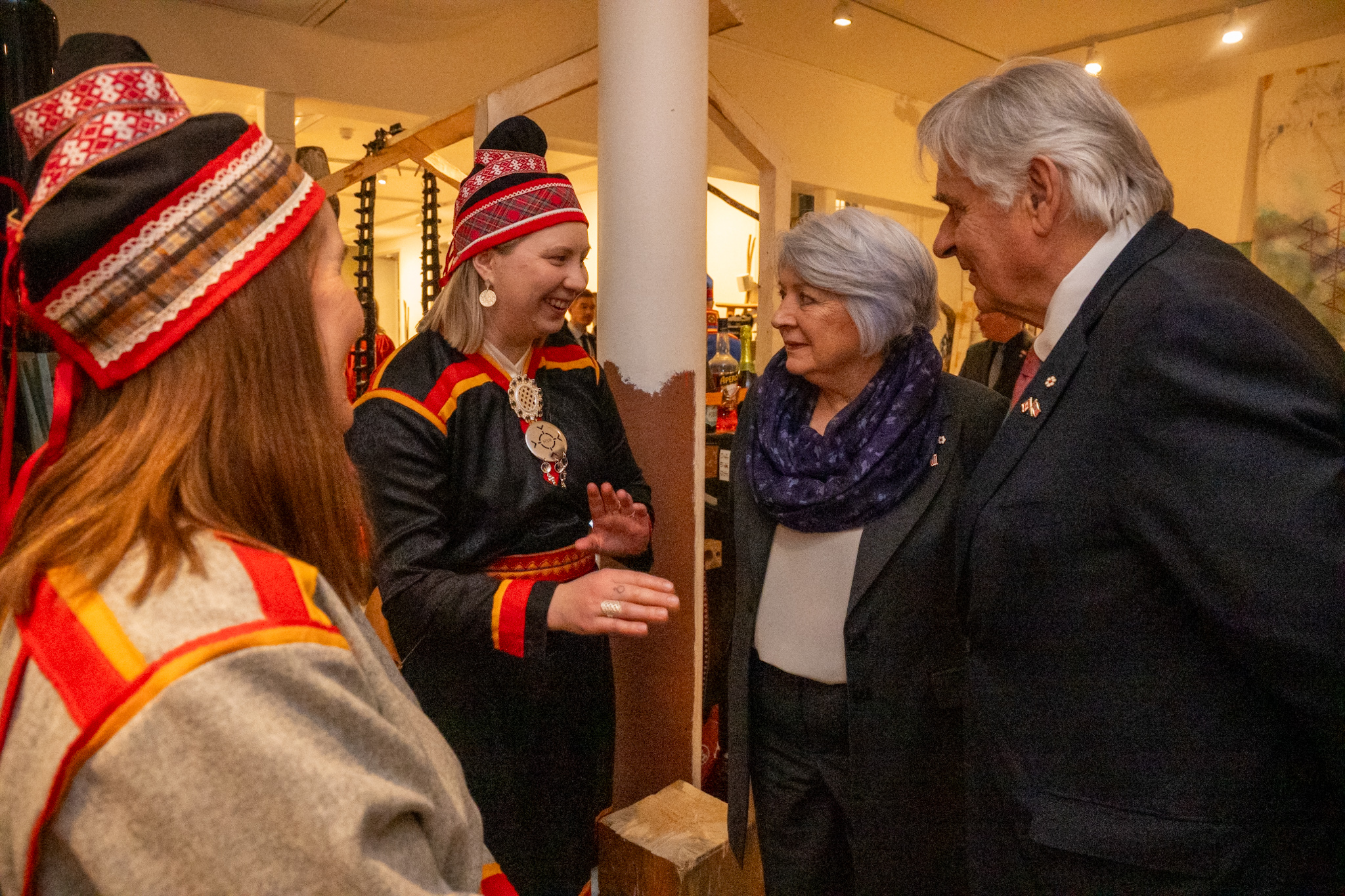 Governor General Mary Simon and Mr. Whit Fraser speaking with two people dressed in traditional Sámi attire with red and yellow patterns. 