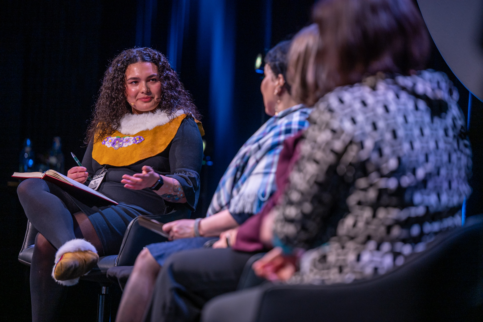 A woman with curly hair in traditional attire leads a panel discussion with three women, all seated on stage, against a dimly lit blue backdrop.