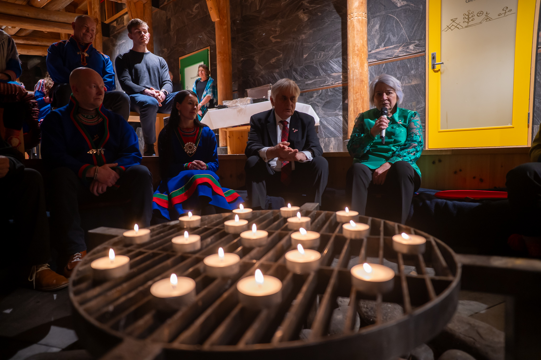 Governor General Mary Simon seated, speaking into a microphone, in a semi-circle of people in a dim, candle lit room.