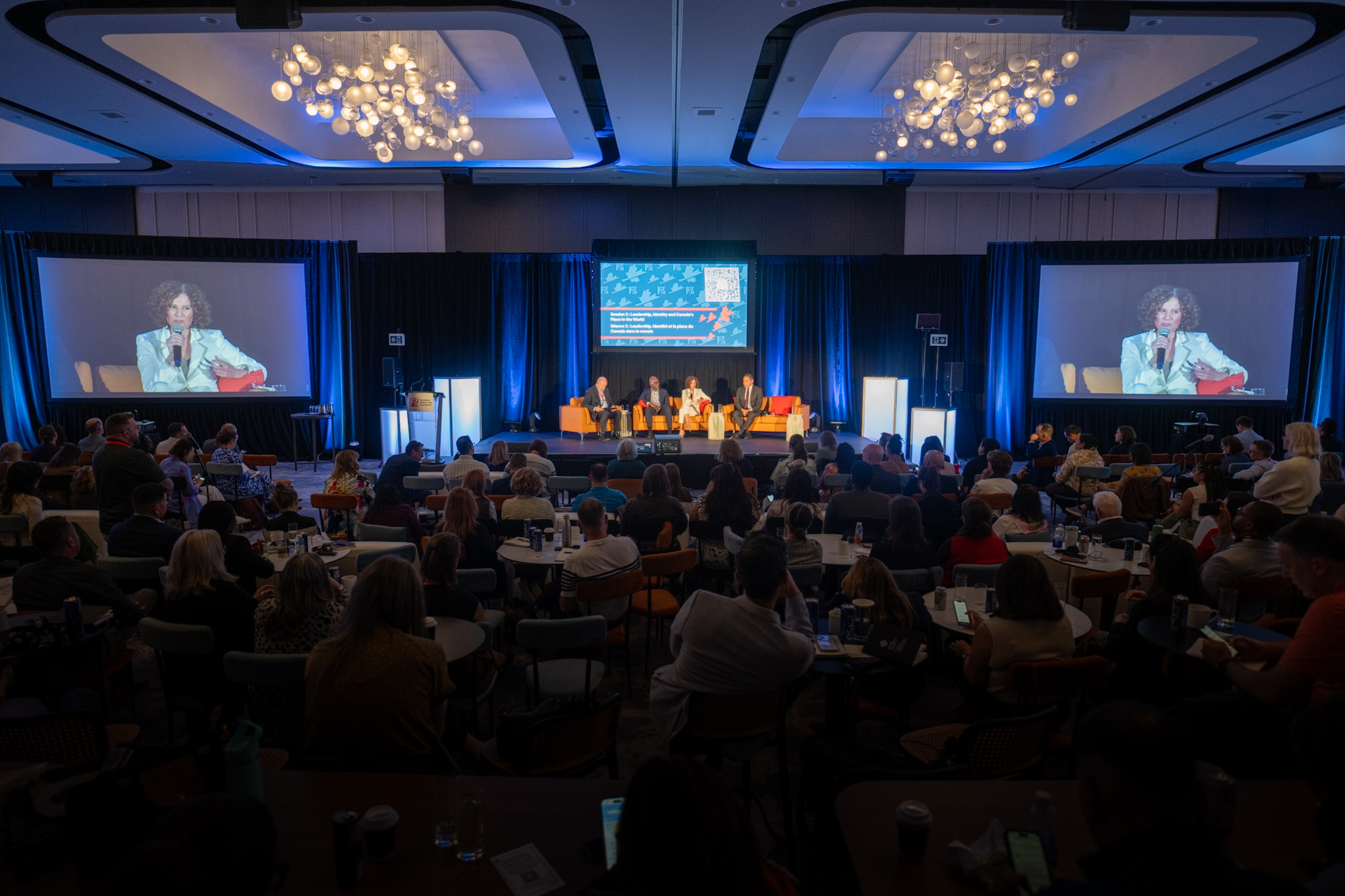 People sitting in a dark room around circular tables, watching a panel of four people onstage.