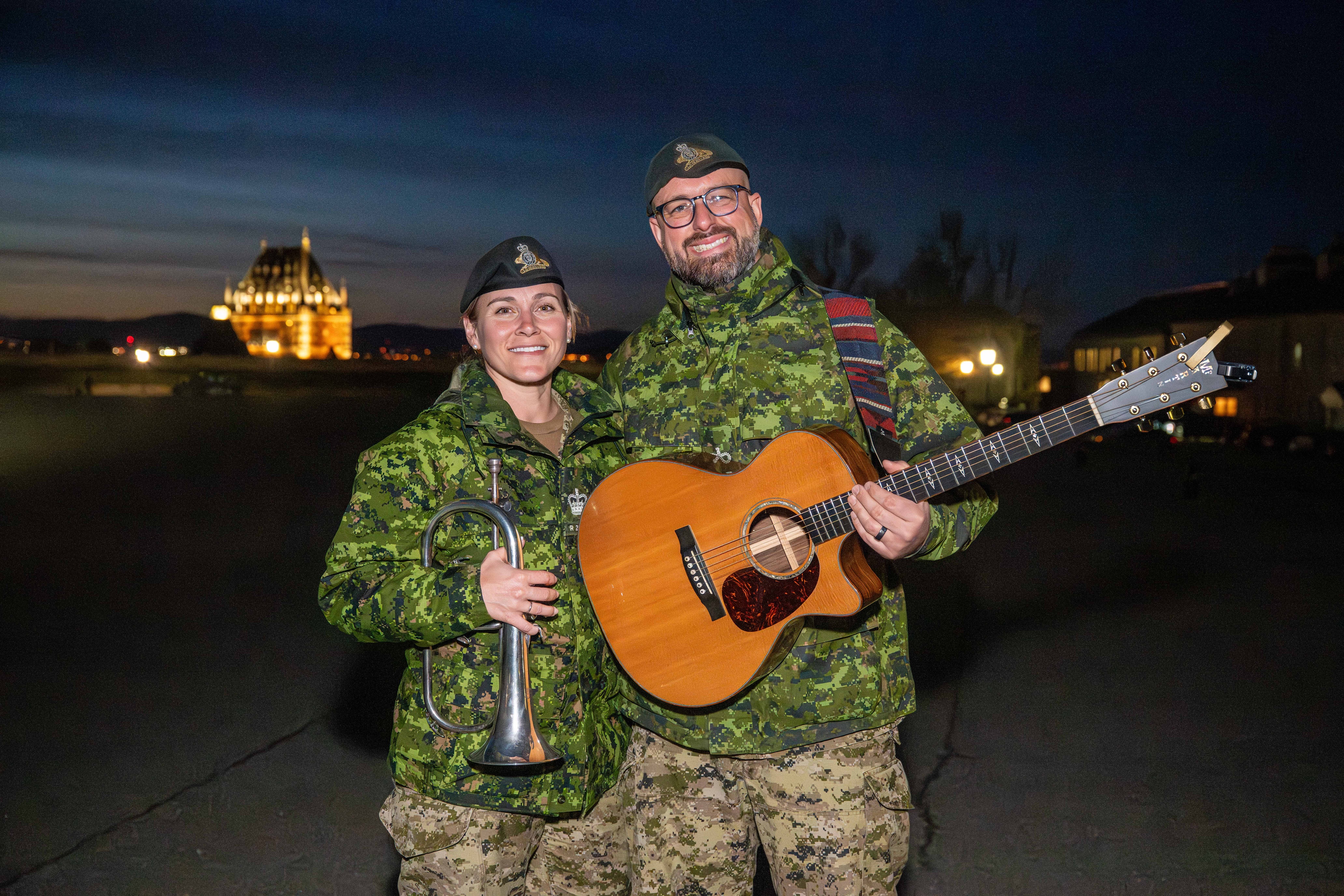 Two people dressed in fatigues are holding instruments, smiling at the camera. It is night time. The Chateau Frontenac is shining behind them. 