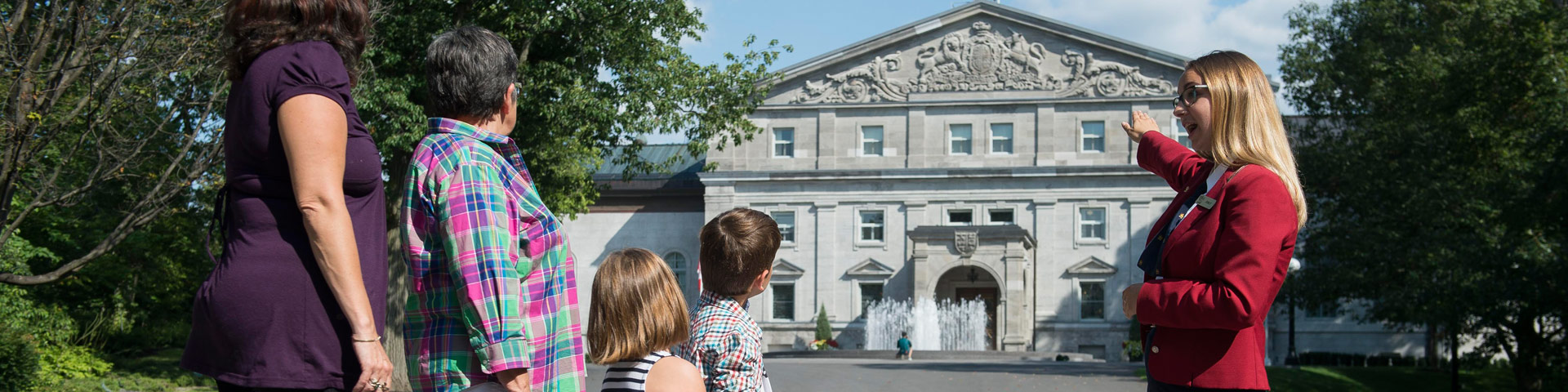 Une guide-interprète montrant la façade de la résidence de Rideau Hall (et une fontaine)  à un petit groupe de visiteurs.