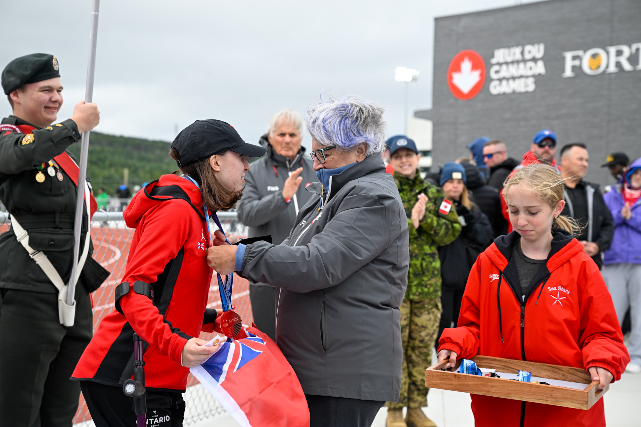 Governor General Mary Simon placing a medal around a woman's neck. A young girl is holding a tray of medals.