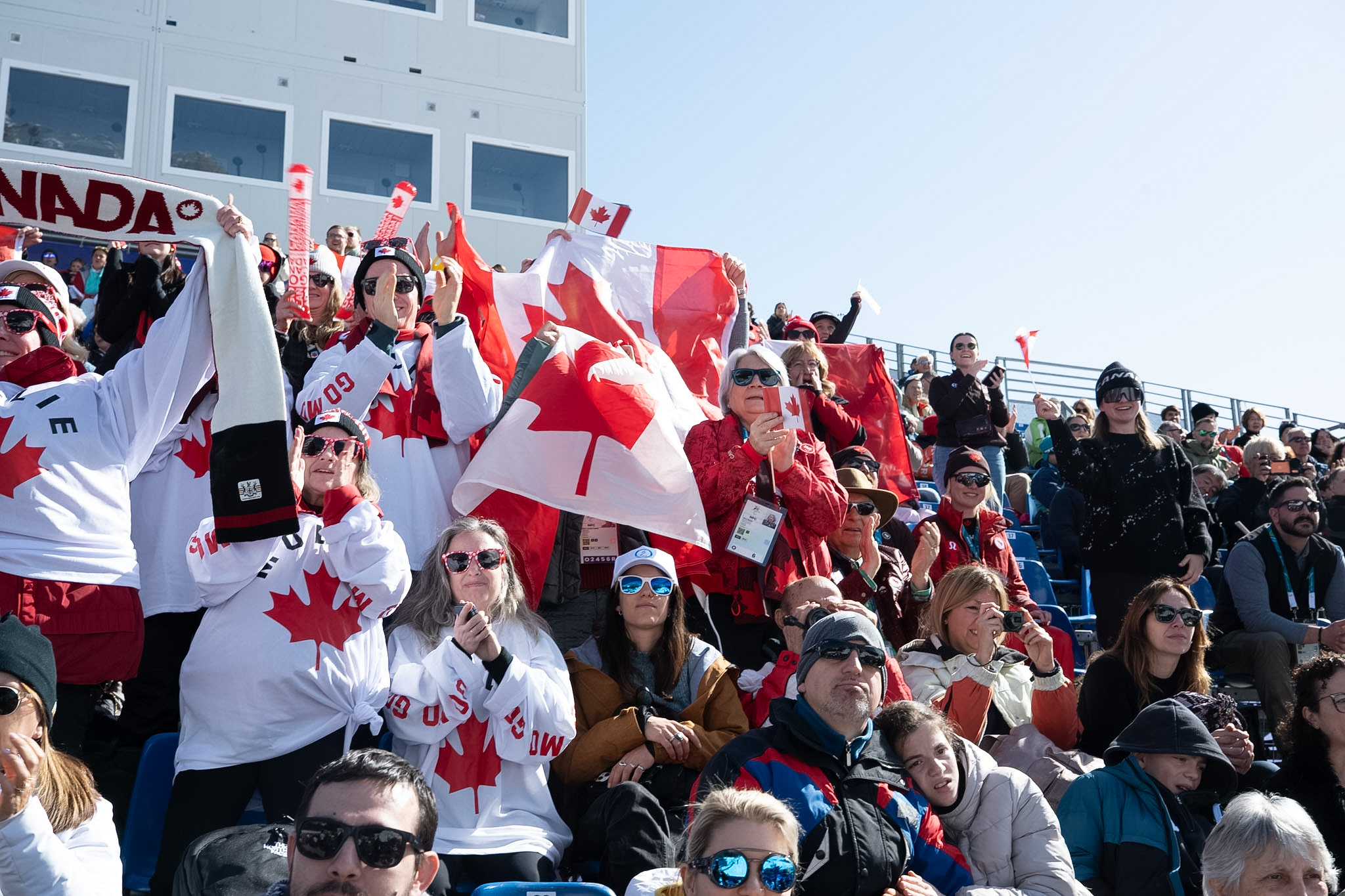 Governor General Mary Simon standing and cheering with a large crowd. Many people are wearing clothing featuring the Maple Leaf and holding Canada flags.