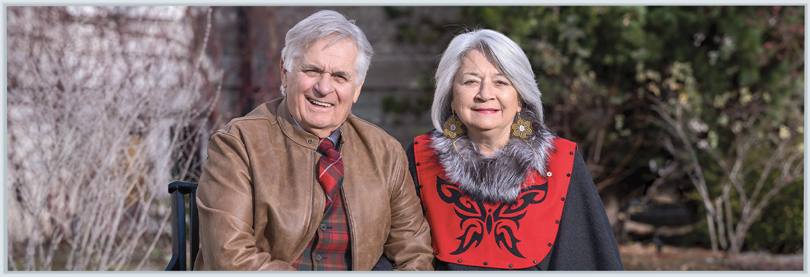Mr. Whit Fraser and Governor General Mary Simon are sitting on a park bench, outside, smiling and holding hands, on a sunny fall day.