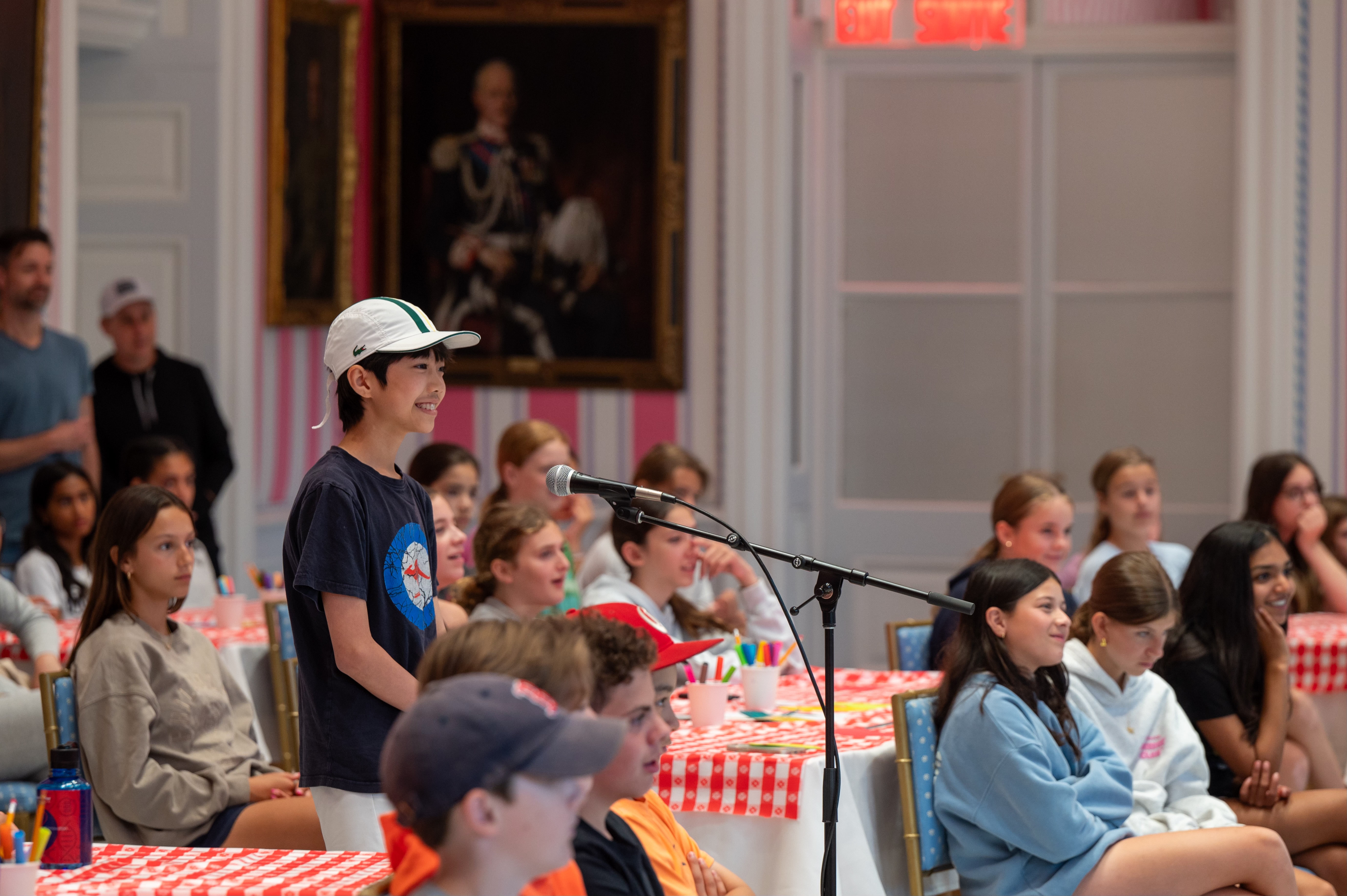 A young student stands at a microphone. He is surrounded by other students and is wearing a ball cap.