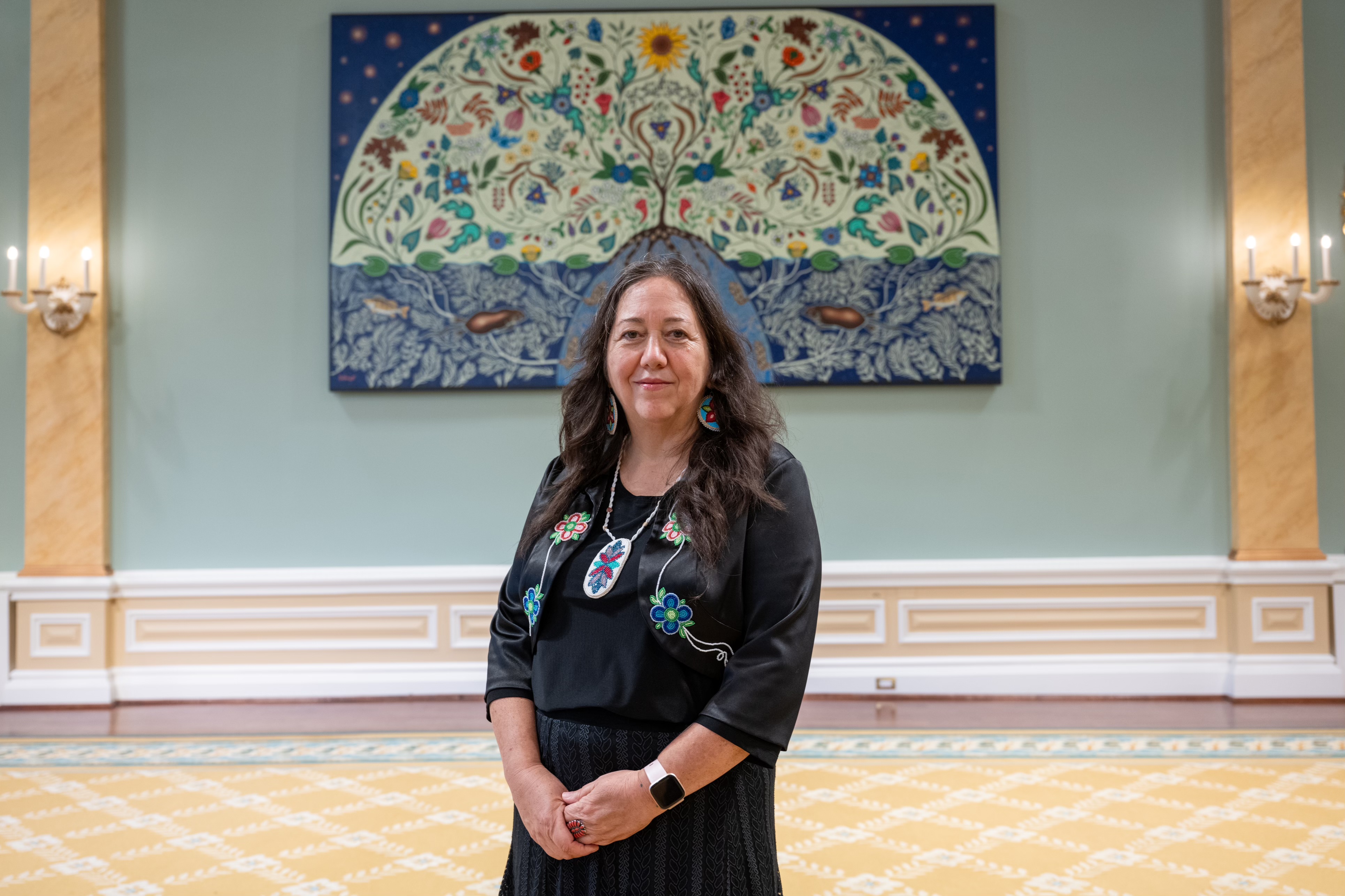 A woman is standing, posing for a photo. There is a large painting on the wall behind her. There is a grey box in the bottom left corner of the photo.