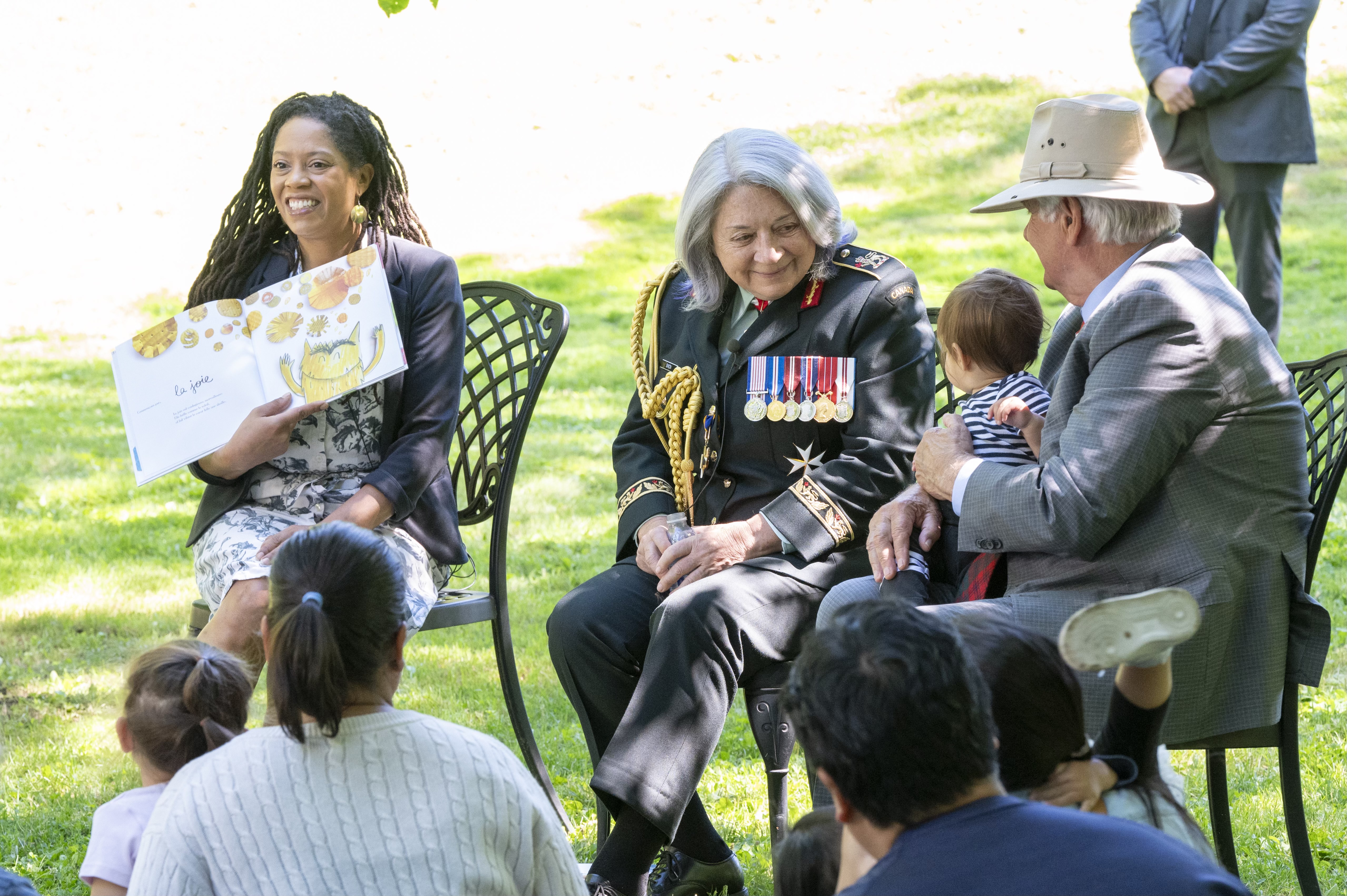 Governor General Mary Simon smiles at a child. She is dressed in the Canadian Army uniform. A women sits to her right holding a book and Mr. Whit Fraser is seated to her left. There are children in the foreground.