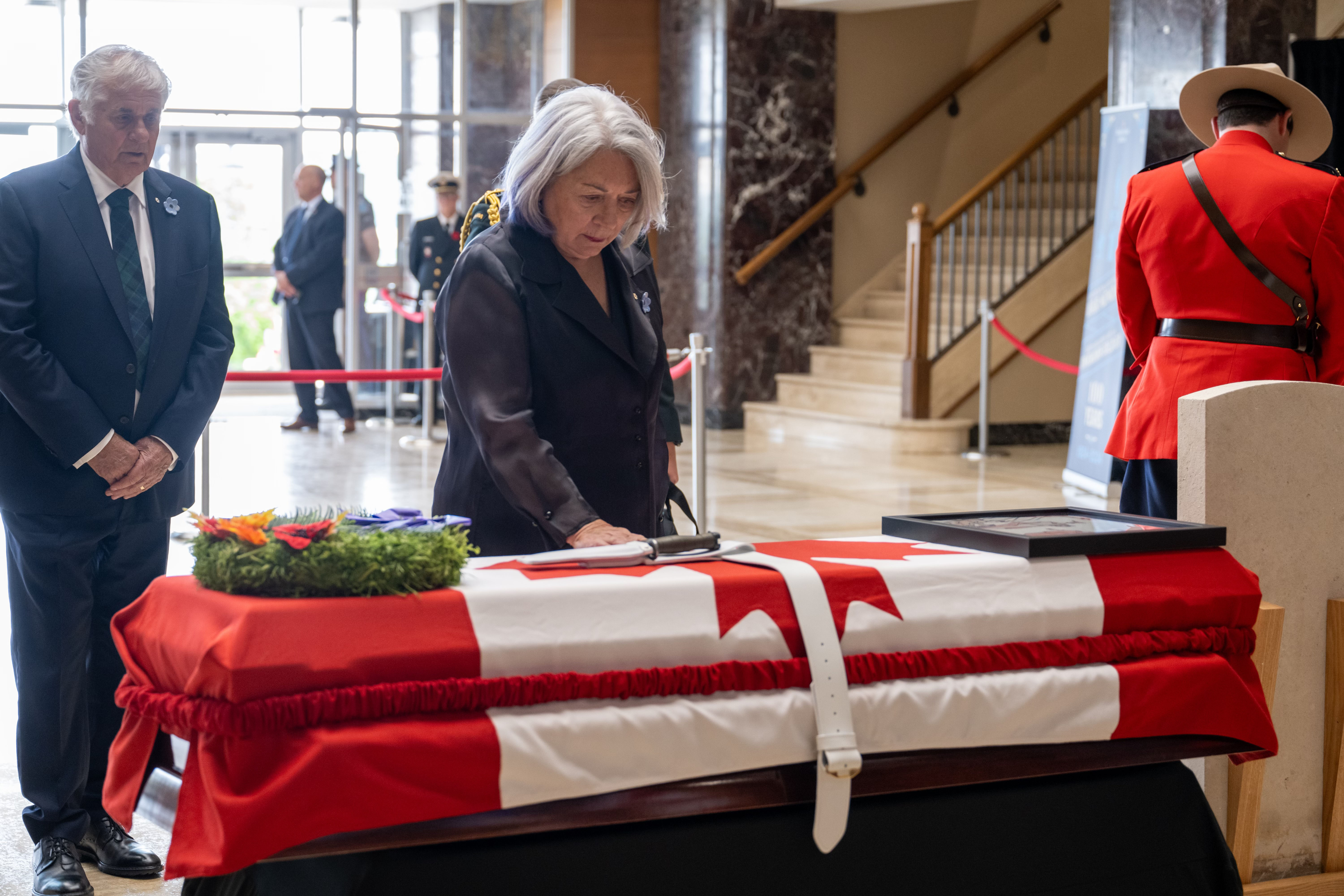 Governor General Simon standing before a casket draped with a Canada flag. There is a wreath and a framed photo on top.