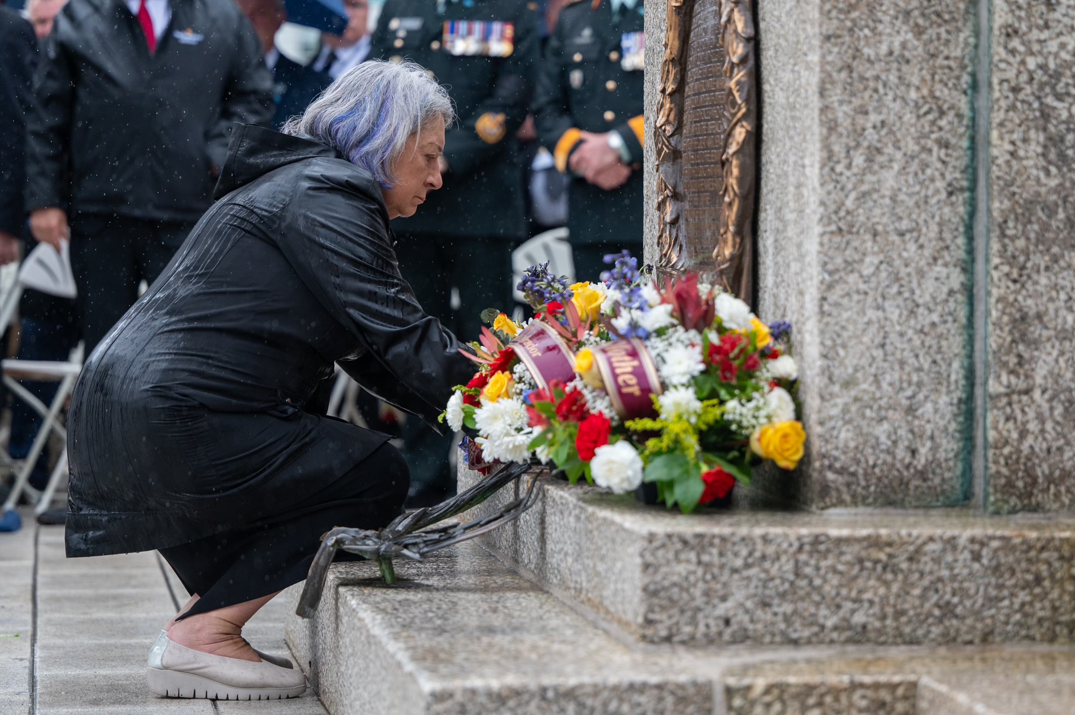 Governor General Simon is crouched down at the foot of a war memorial. There is a plaque on the wall of the memorial and a bouquet of flowers.