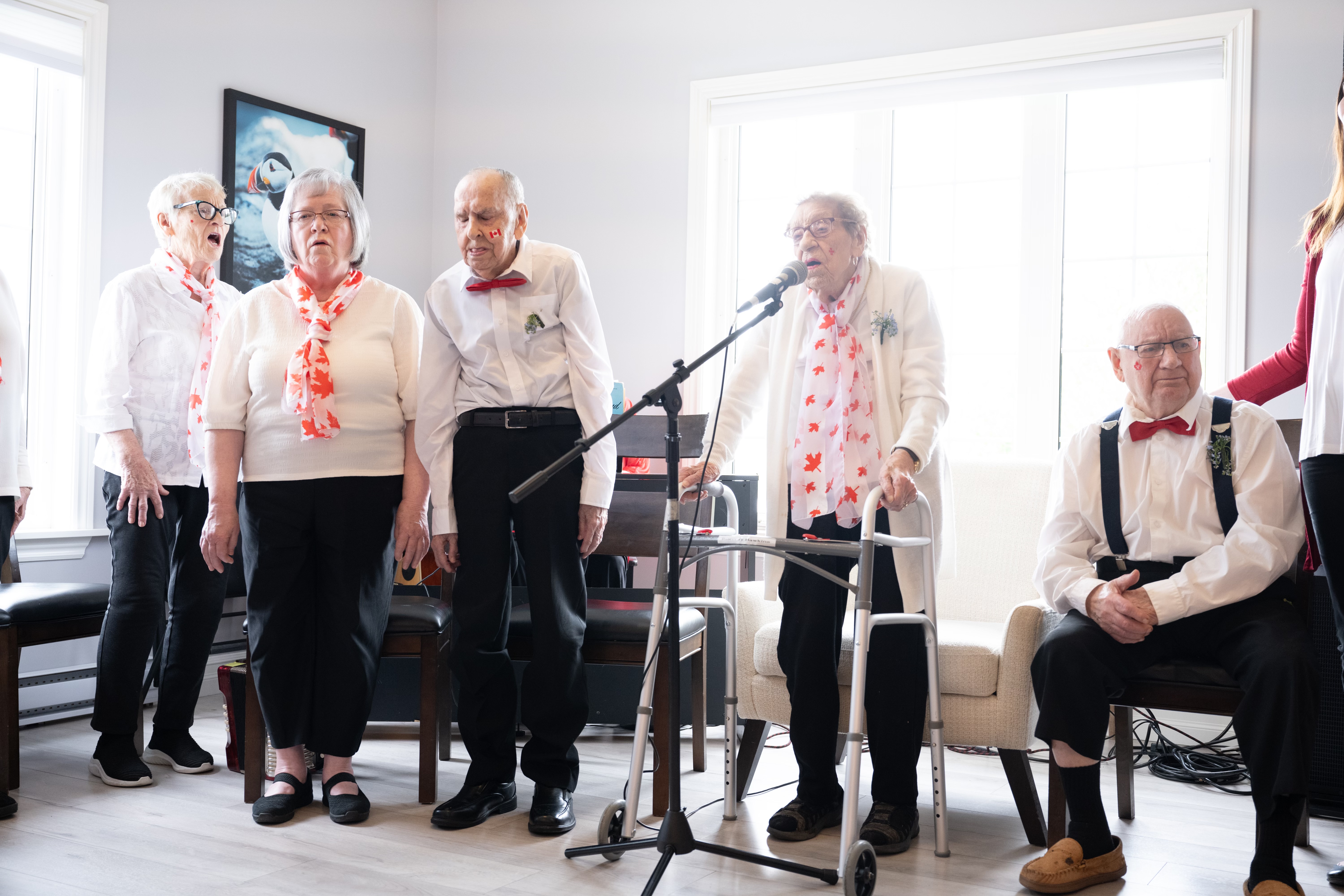 People dressed in red and white for a Canada Day celebration at a retirement home. Four people are standing and singing. There is a microphone in front of one woman who is standing with a walker. One person is seated.
