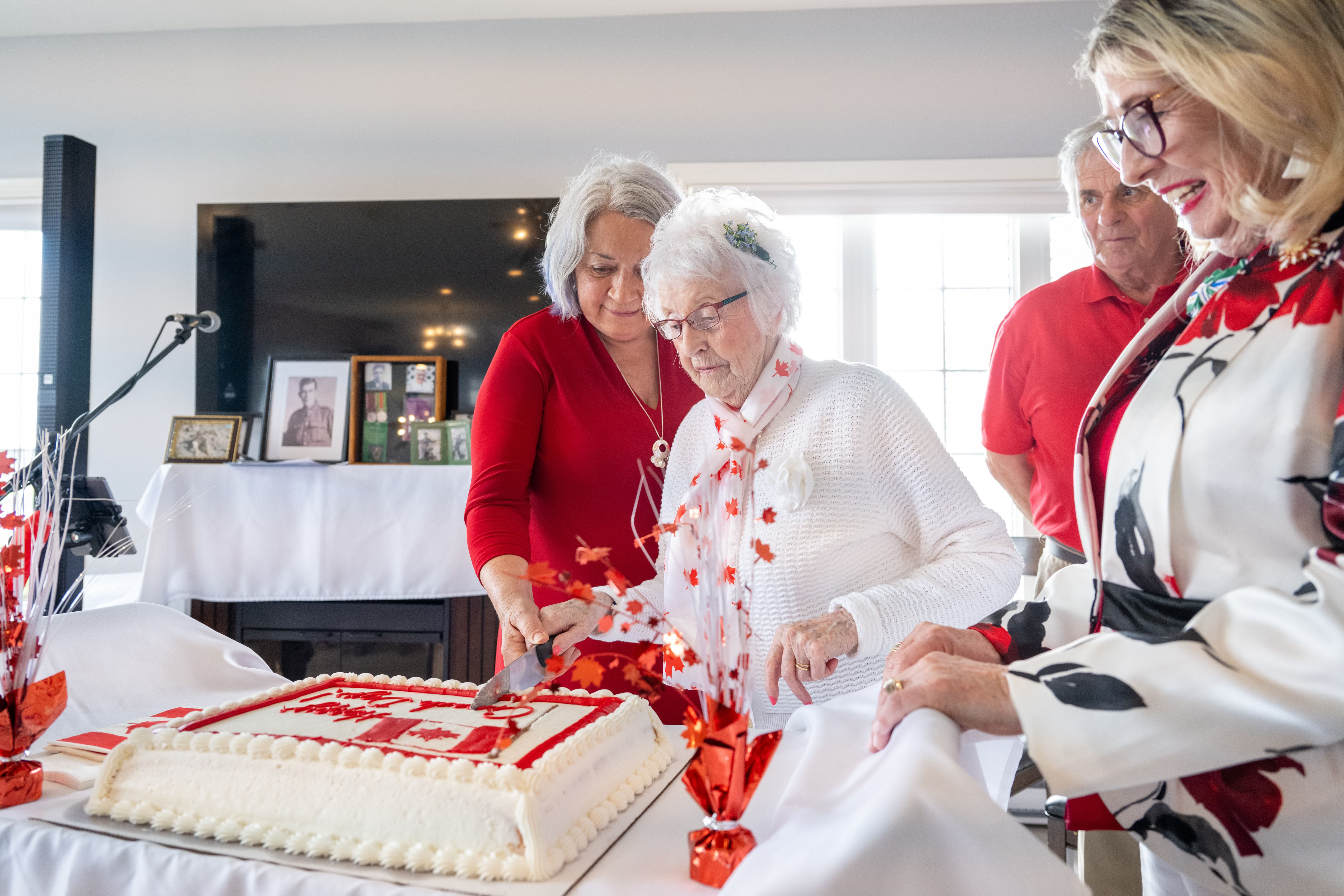 People dressed in red and white for a Canada Day celebration at a retirement home. Governor General Simon is helping a woman slice into a large cake.