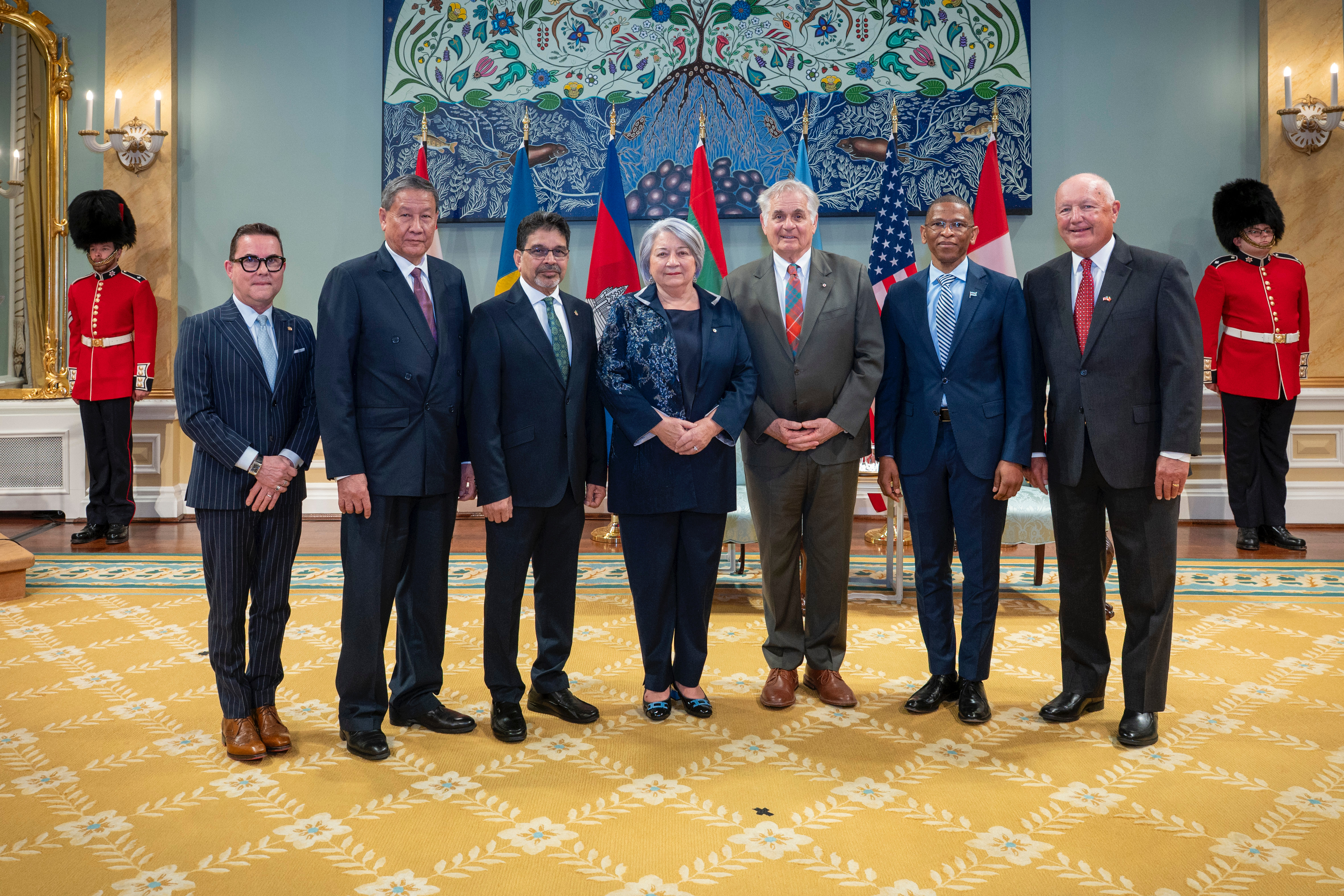 Governor General Simon and Mr. Whit Fraser standing with five new heads of mission, with the flags of each country behind them. Two Ceremonial Guards are in the back.