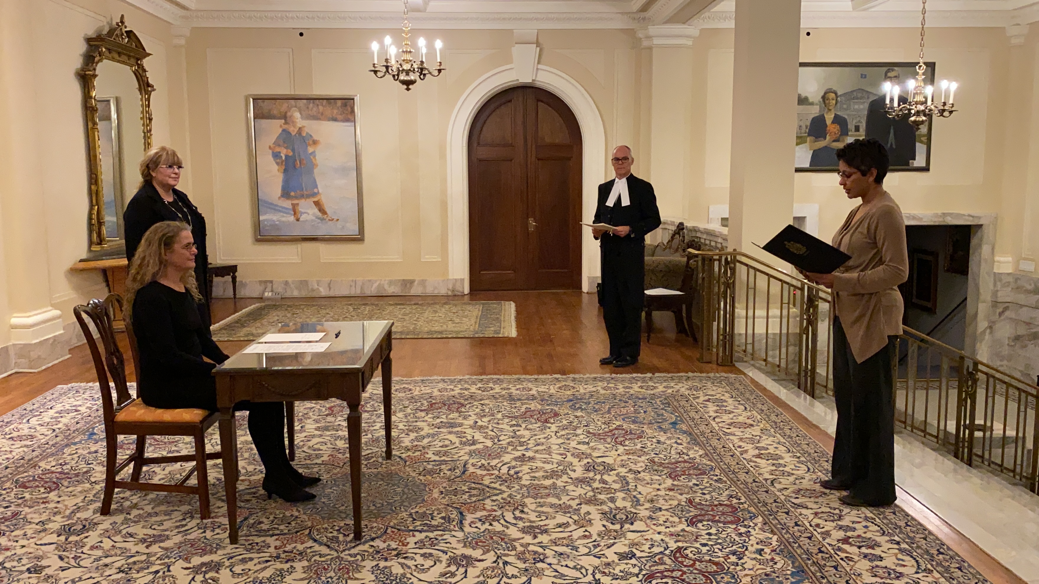 La gouverneure générale est assise à un bureau. Sur le côté droit, il y a un homme qui tient un document dans ses mains. Quatre autres personnes sont debout autour du bureau tout en respectant les normes de distanciation sociale.